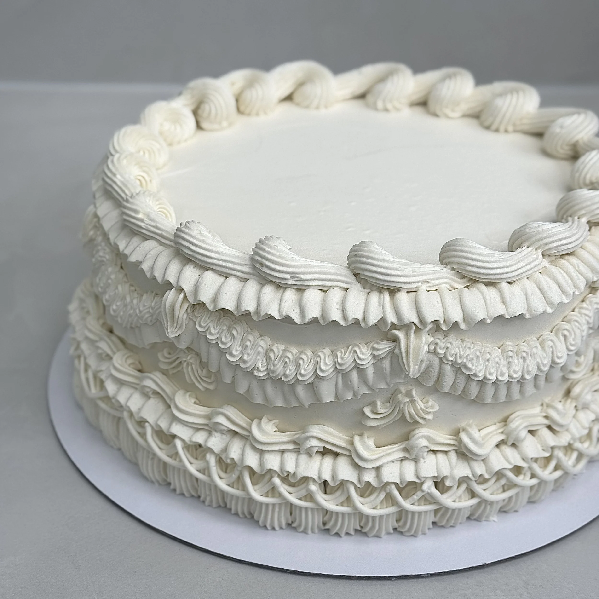 A white, multi-layered cake decorated with piped frosting designs and border embellishments.
