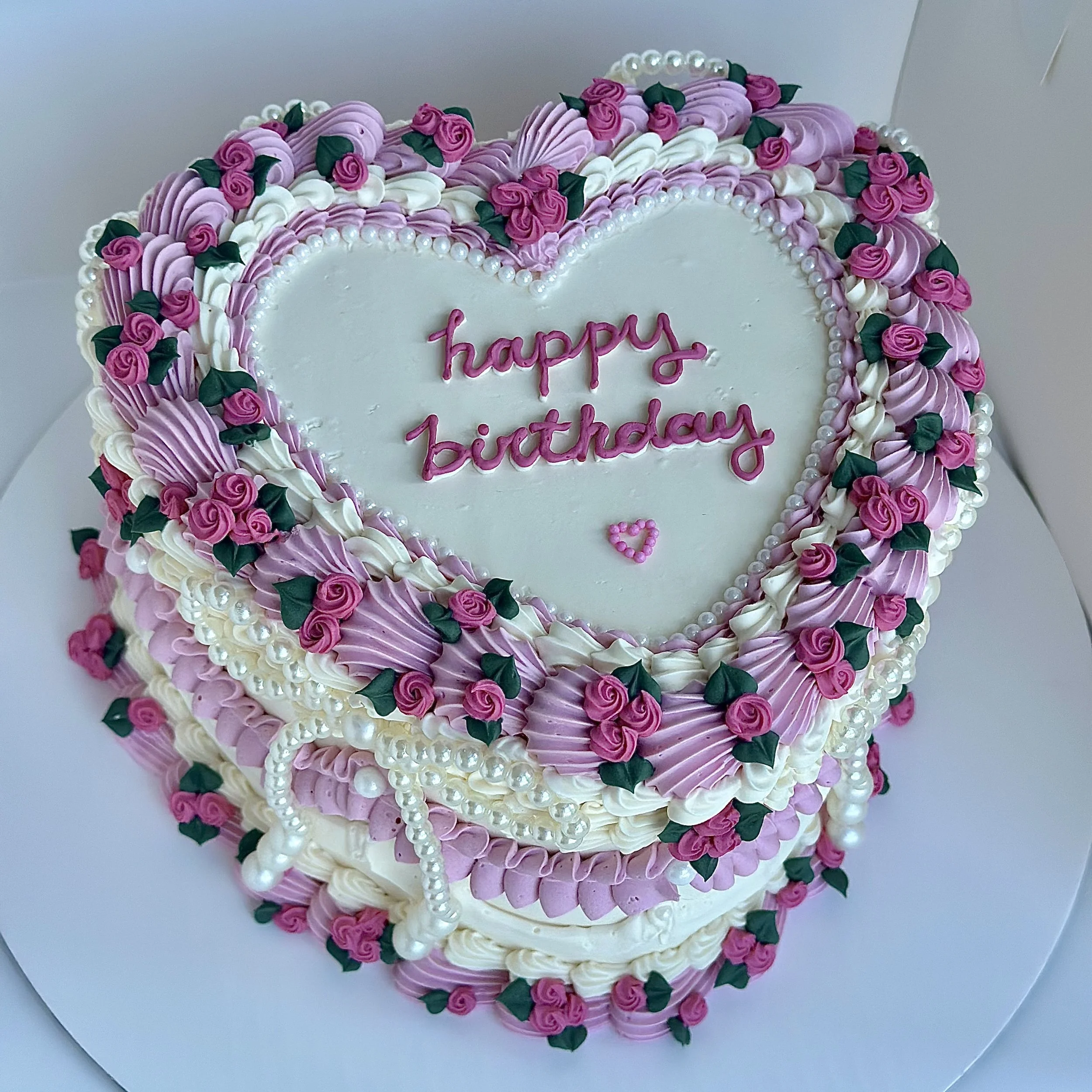 Heart-shaped birthday cake decorated with pink and white frosting, small pink roses with green leaves, pearl-like candies, and a pink message that reads 'happy birthday'