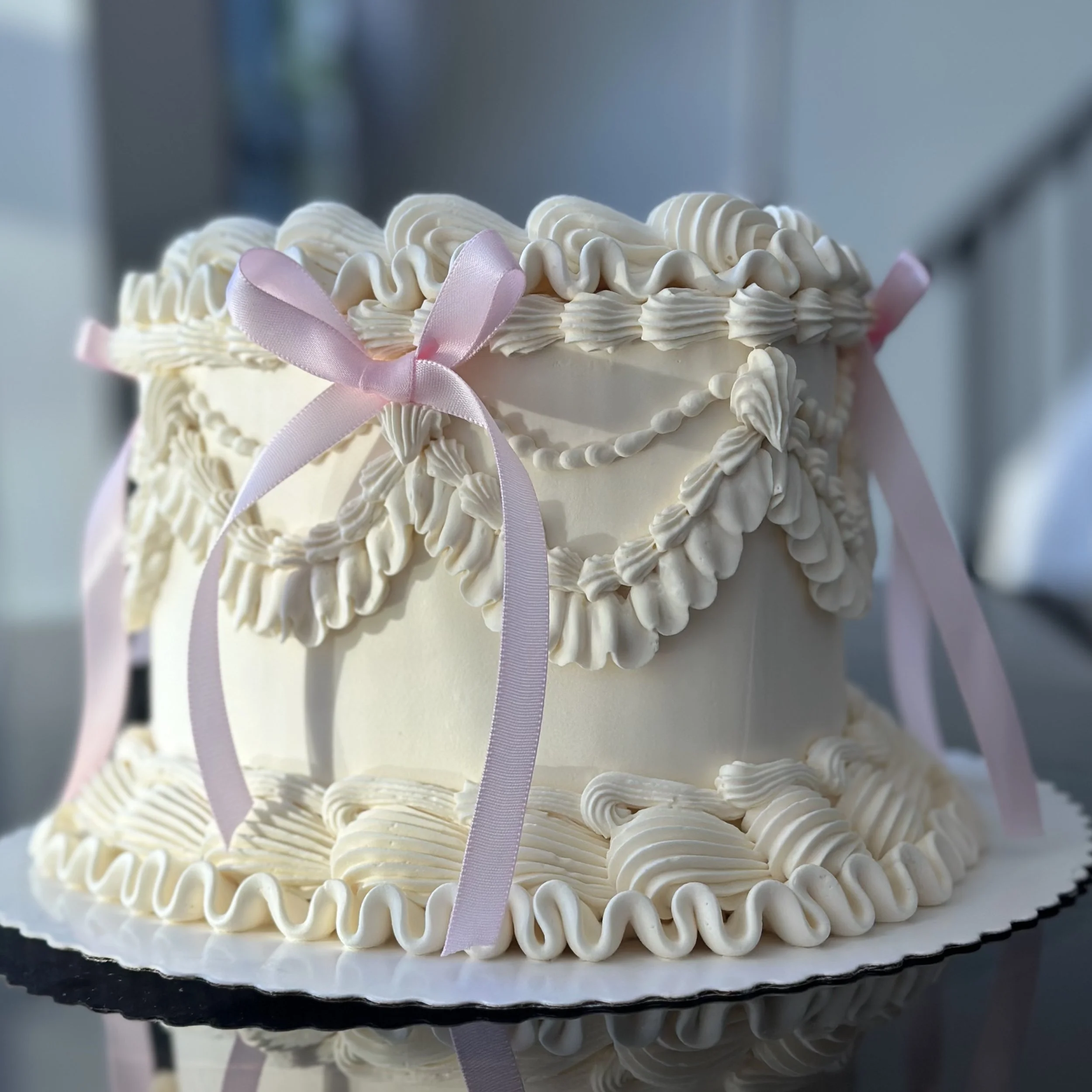 A white birthday cake decorated with piped cream and pink ribbons, featuring intricate design details.
