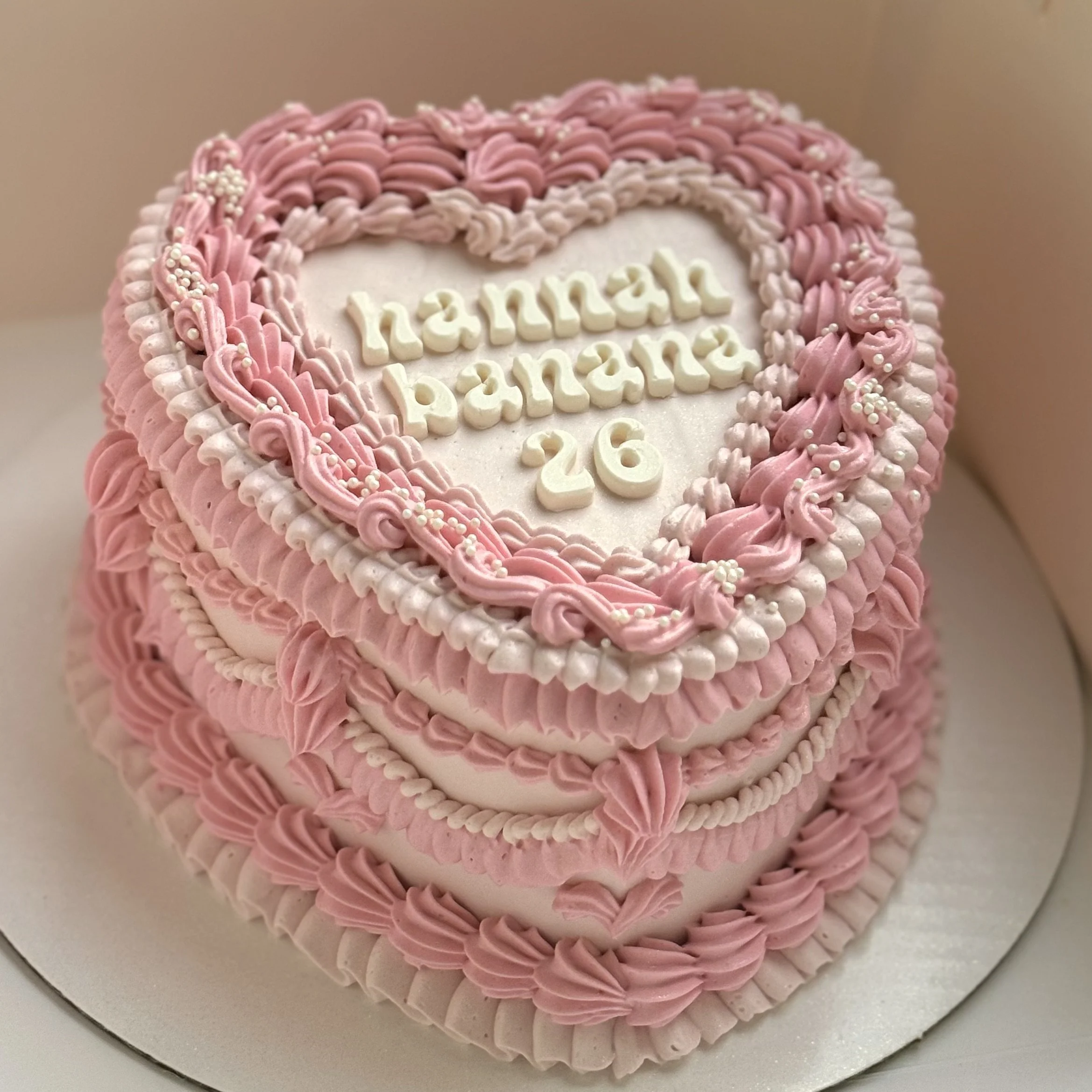 A heart-shaped pink and white birthday cake with pink and white frosting decorations. The cake has the message "happy birthday 26" written on it. The cake is placed on a silver cake board.