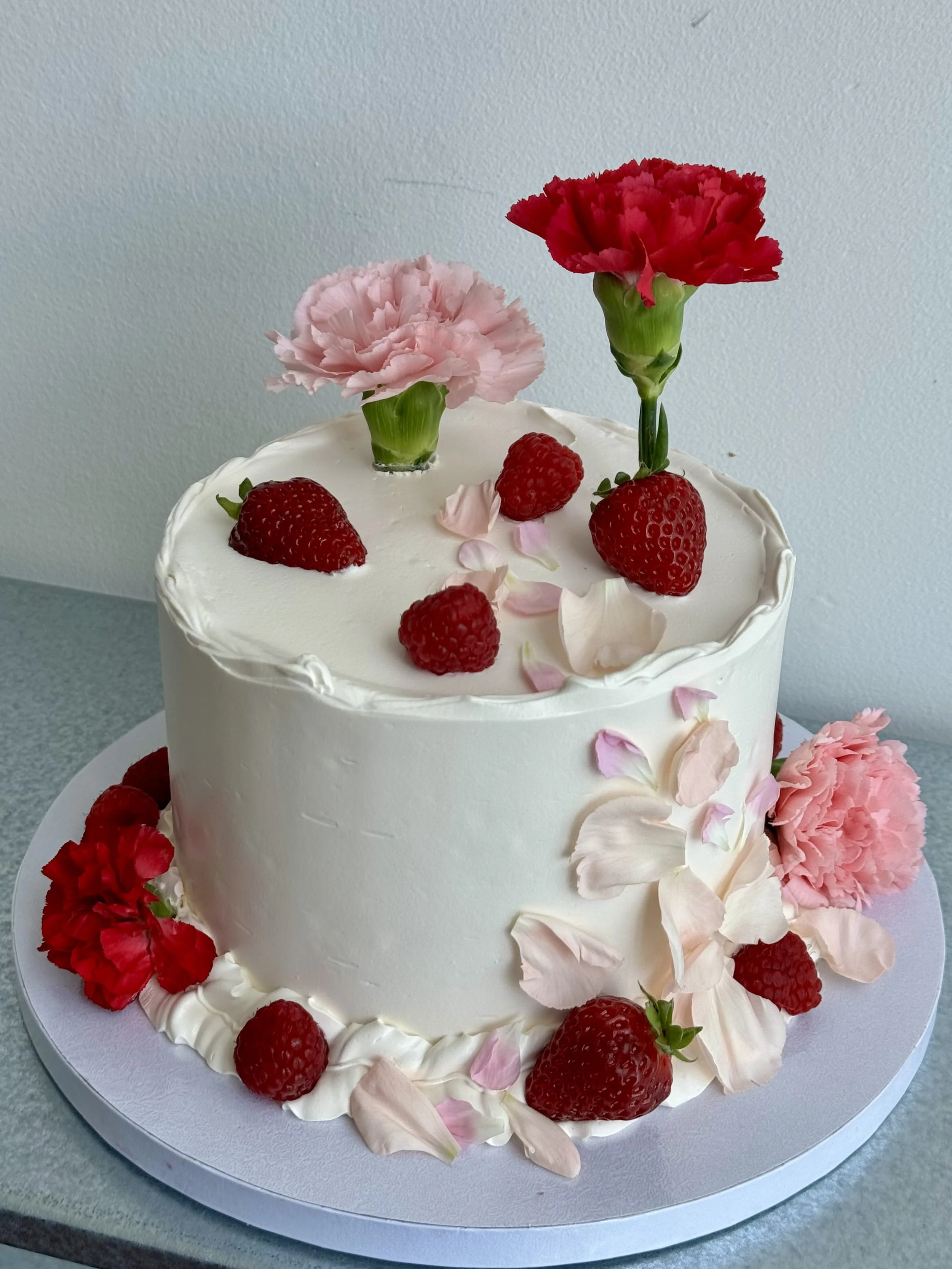 A white frosted cake decorated with pink and red carnations, strawberries, pink rose petals, and cream piping on the base.