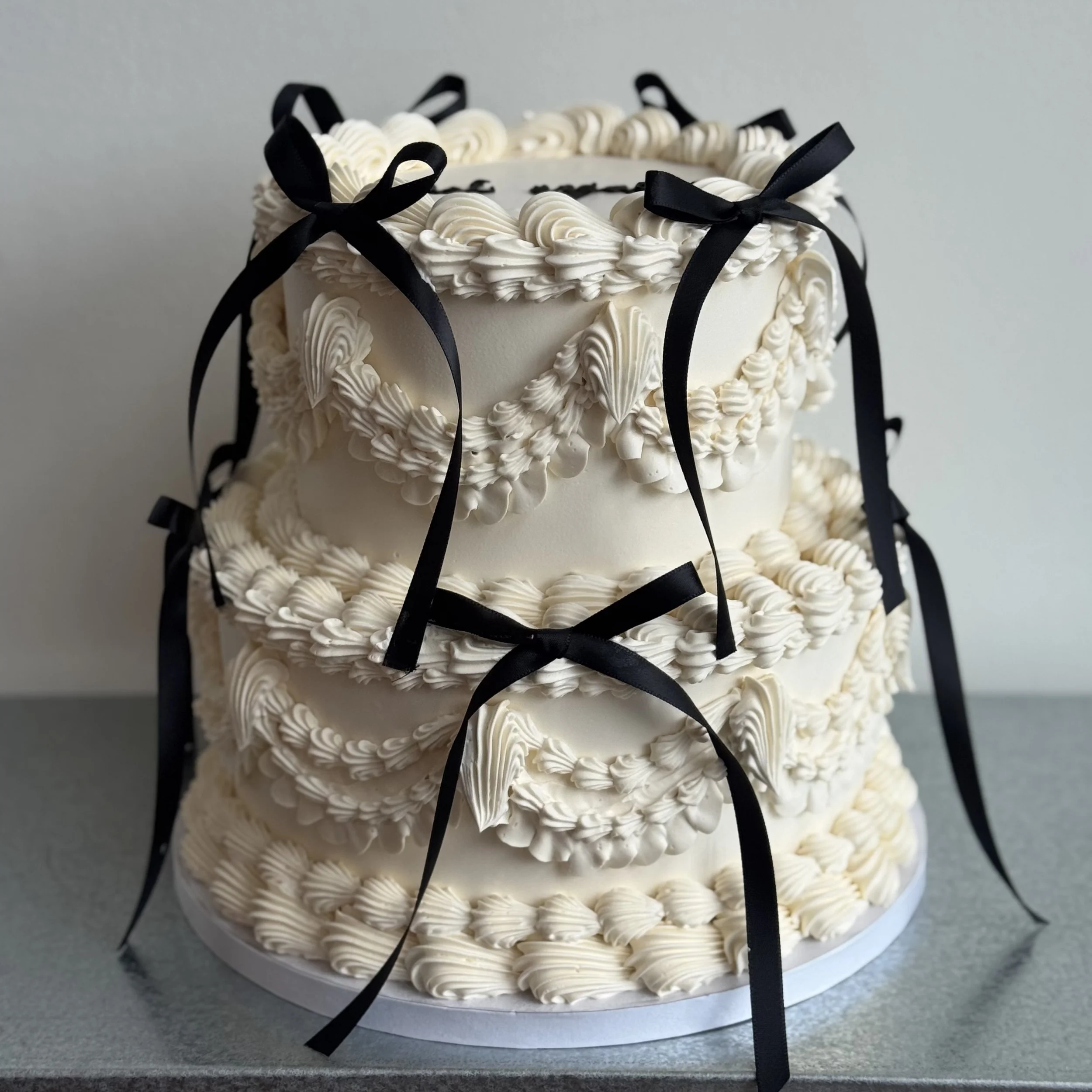 A three-tiered white wedding cake decorated with piped icing and black ribbons tied into bows.