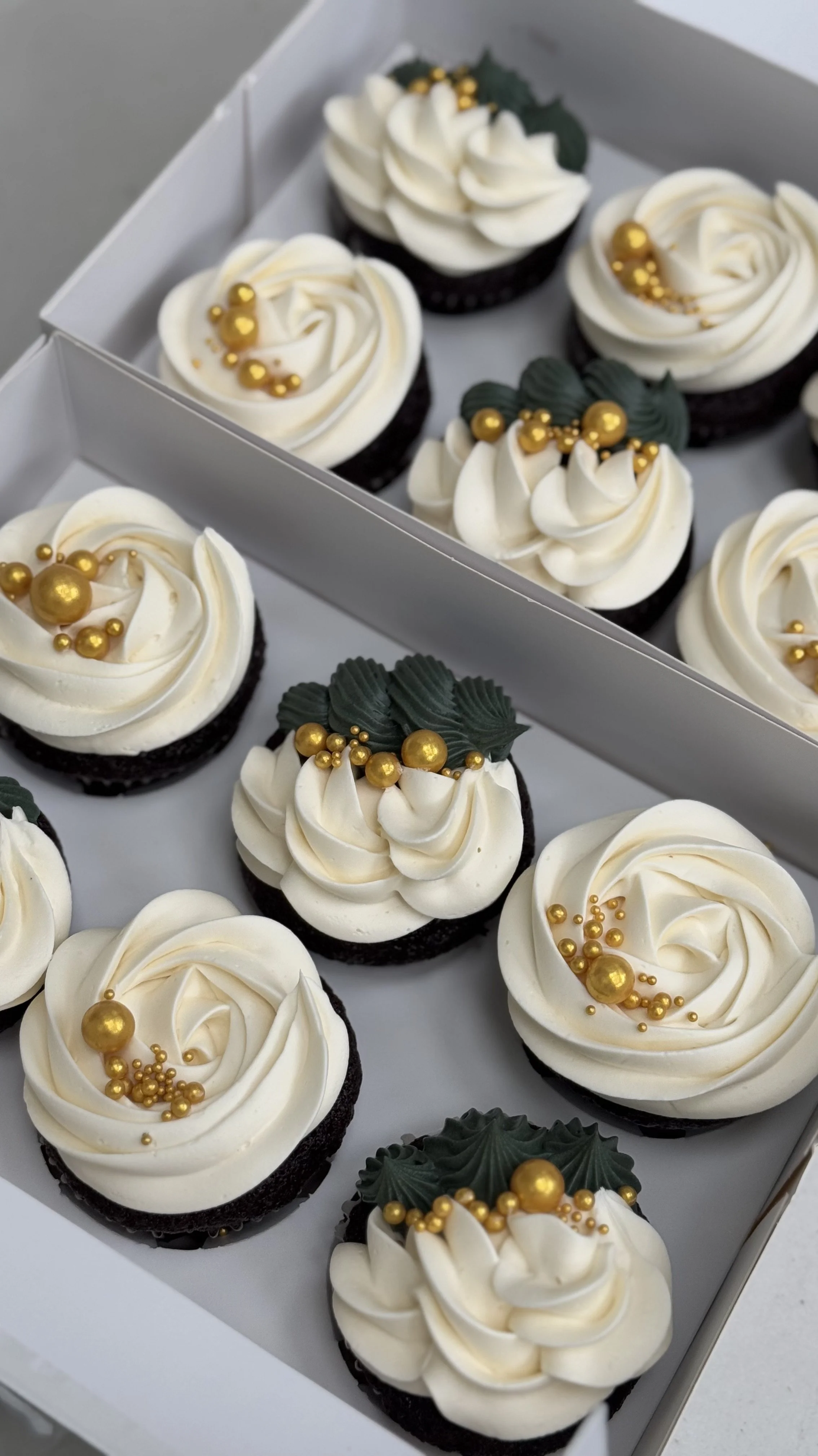 A box of decorated cupcakes with white frosting, gold edible pearls, and green leaf-shaped decorations.