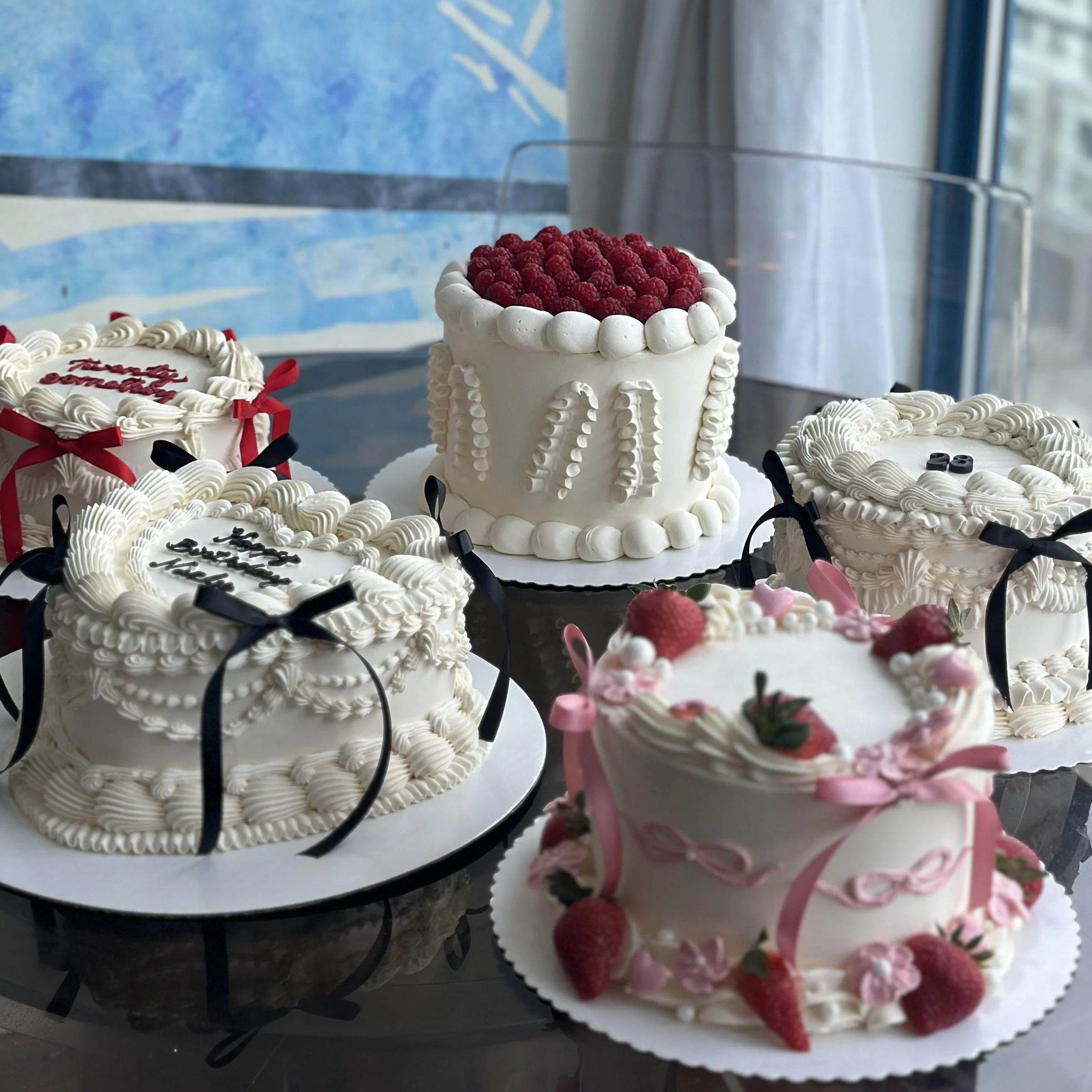 Five decorated cakes with various designs, including strawberries, whipped cream, ribbons, and messages, on a table near a window.