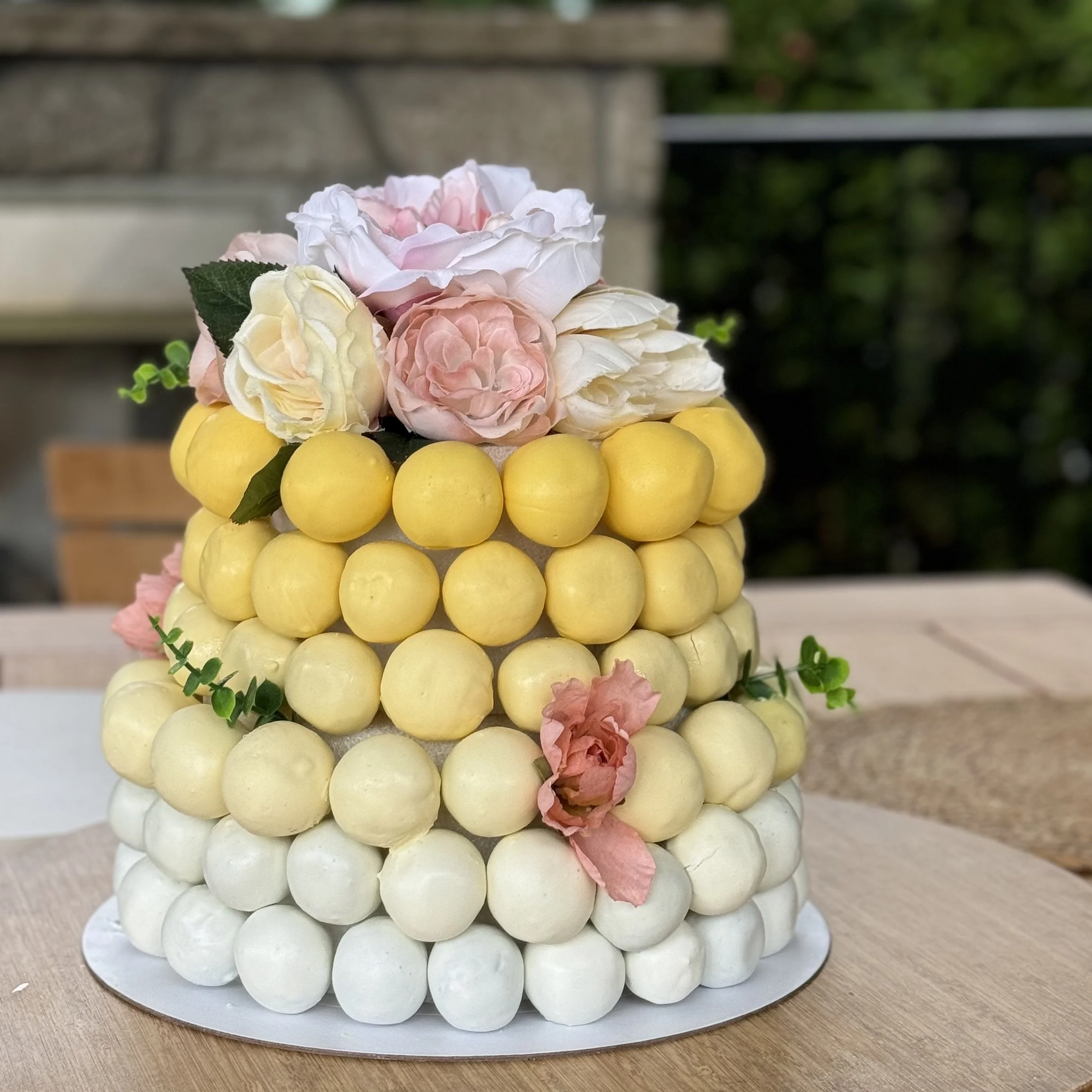 A multi-tiered cake decorated with yellow, white, and pink armature spheres topped with pink and white flowers and green leaves, set on a wooden table outdoors.