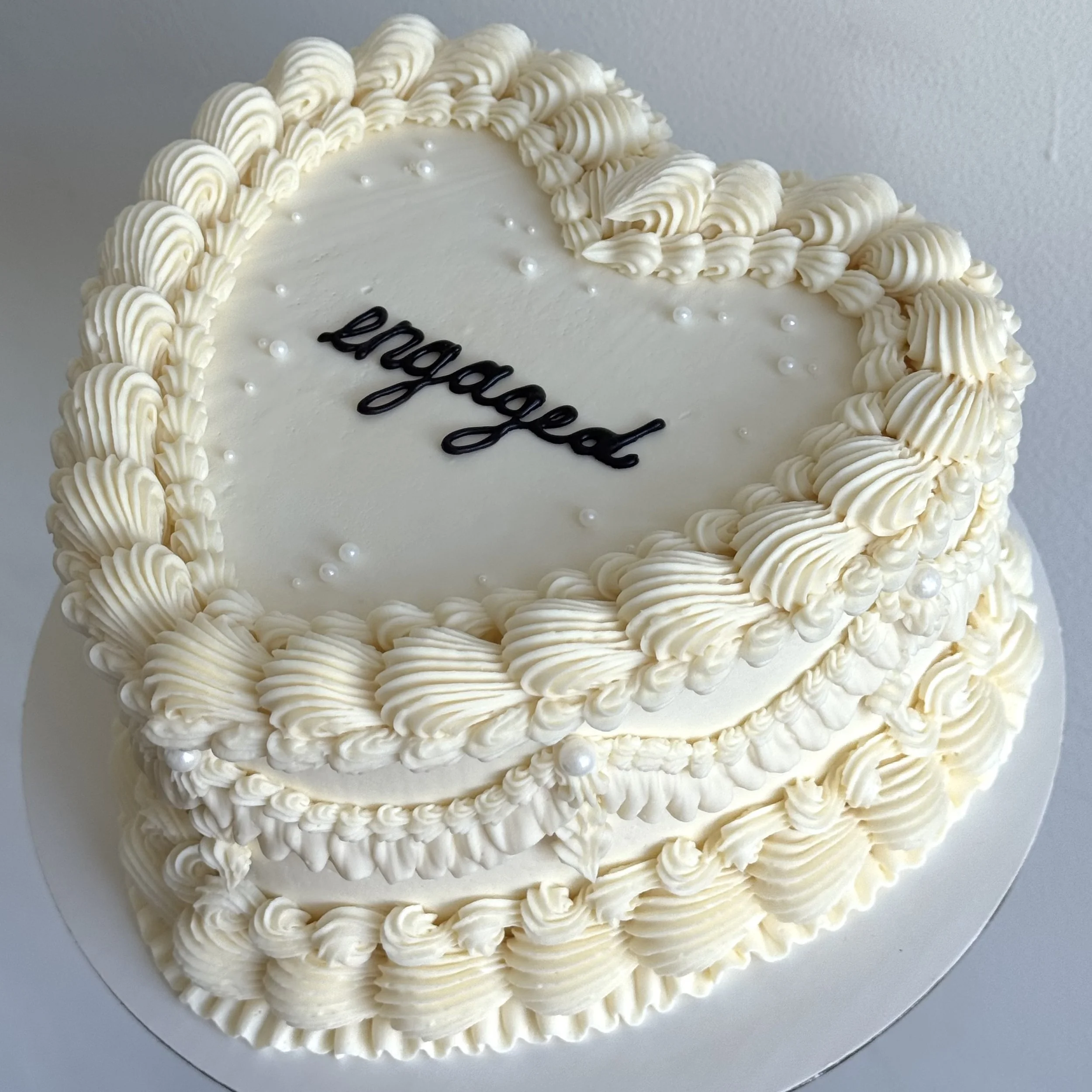 Heart-shaped white cake decorated with piped cream border, small white pearl-like balls, and the word "beloved" written in black icing on top.