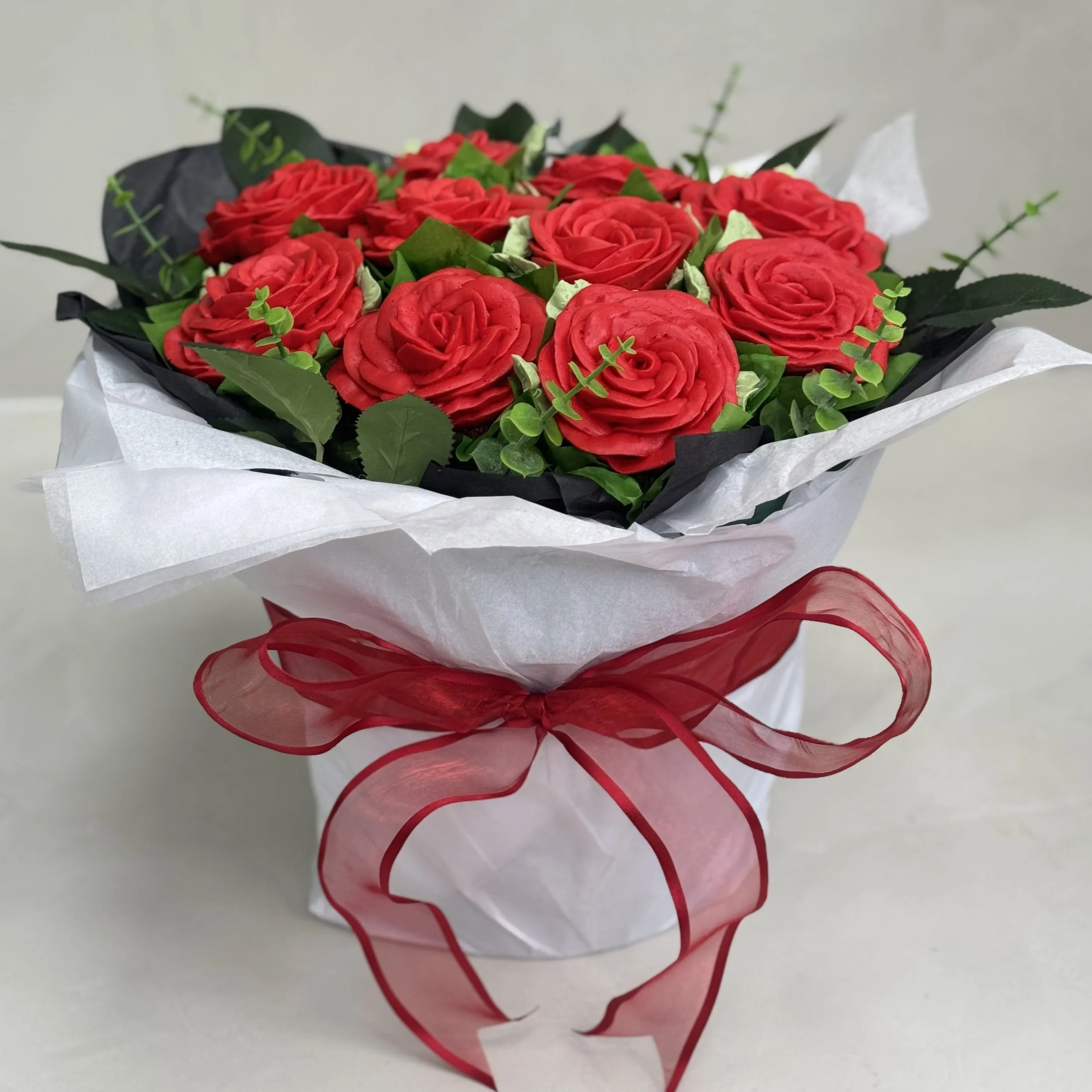 A bouquet of red roses with green leaves wrapped in white paper and tied with a red ribbon.
