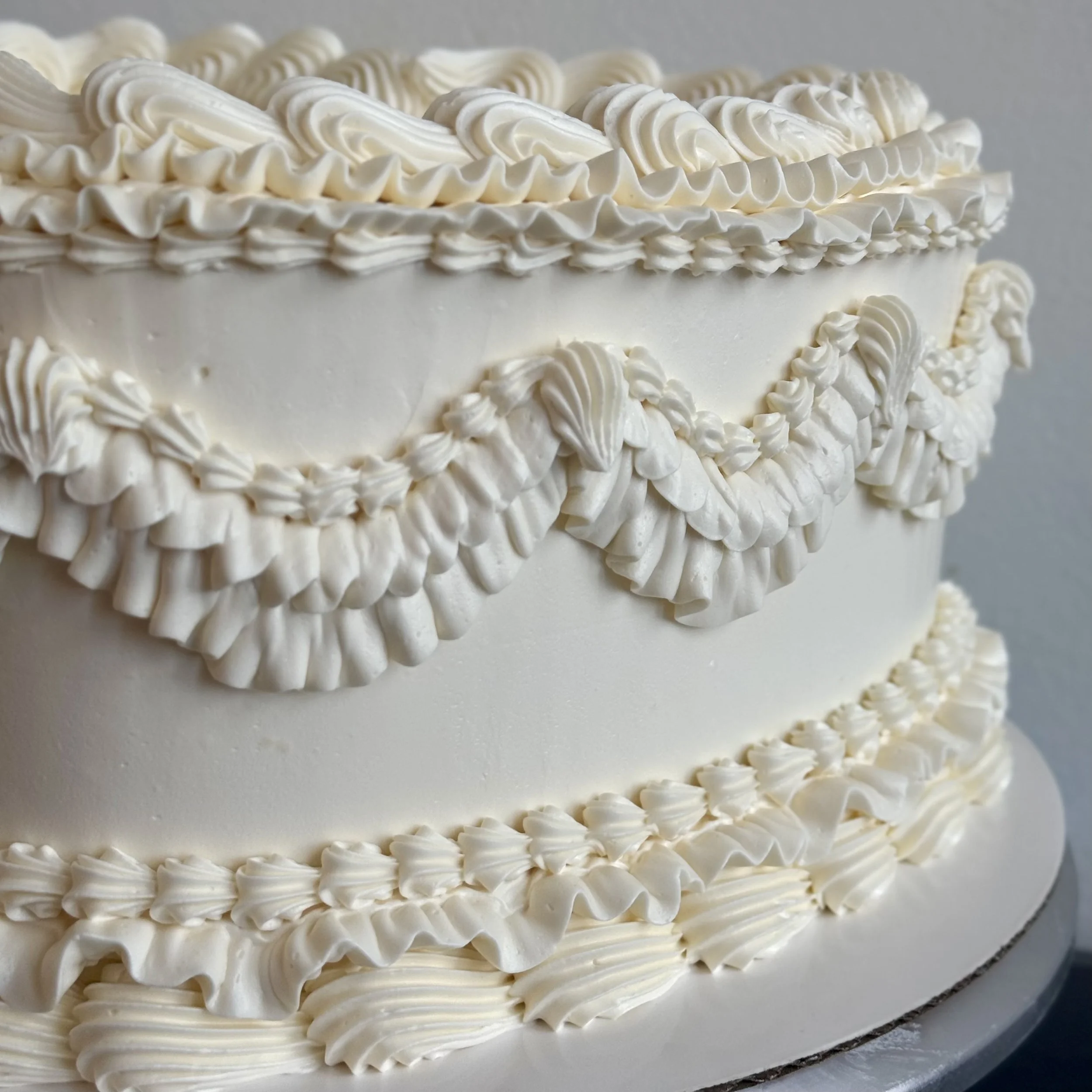 A close-up of a white tiered wedding cake decorated with piped white frosting in floral and scroll patterns.