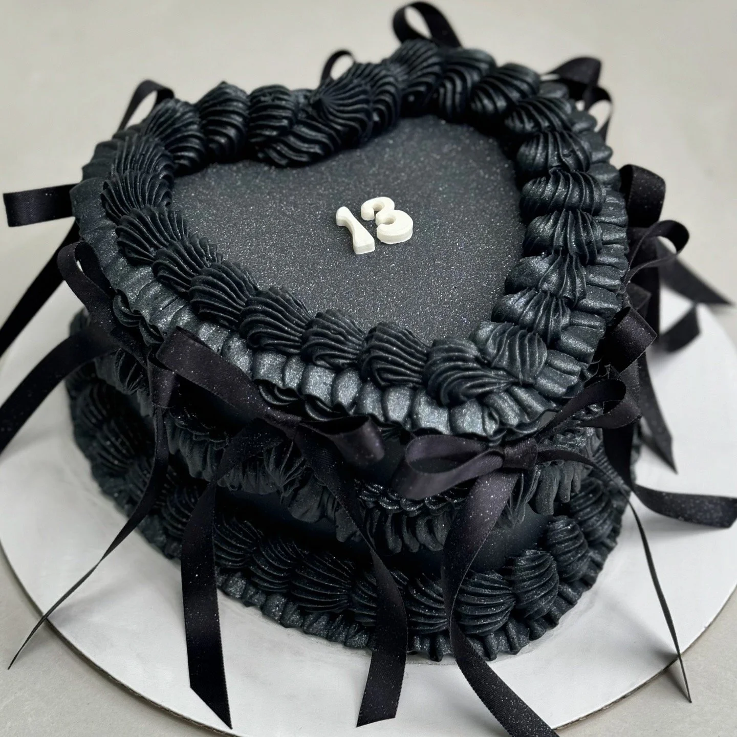 A black, heart-shaped layered cake decorated with piped black frosting, black ribbons, and the number '13' on top.