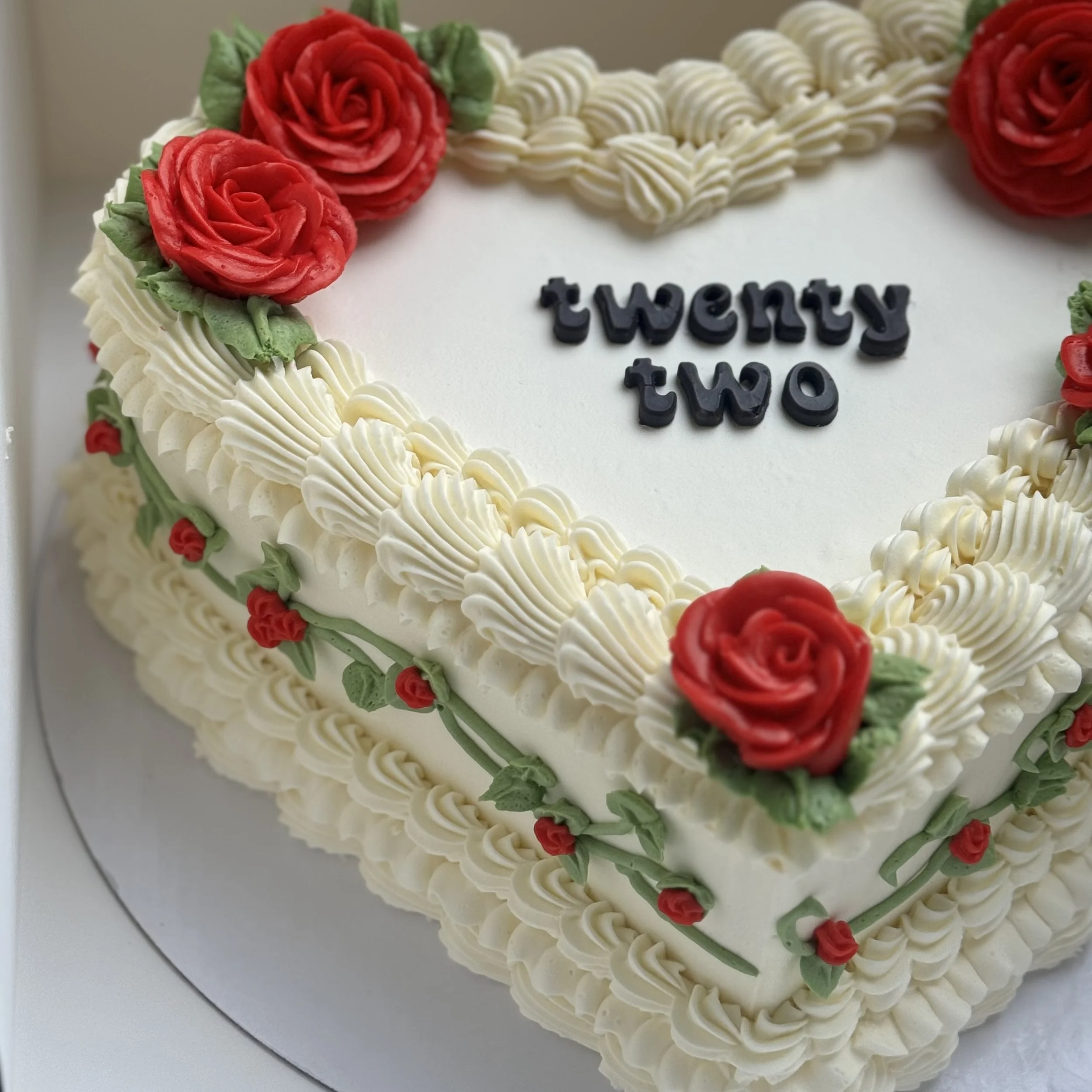 A decorated heart-shaped birthday cake with white frosting, red roses, and green leaves. The cake has the number twenty two written in black icing on top.