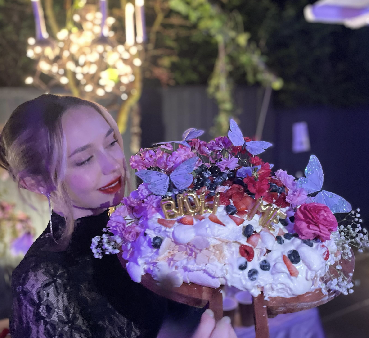 A woman with blonde hair and red lipstick holding a large, decorated birthday cake with pink and purple flowers, blue butterflies, and strawberries it has a gold 'LOVE' decoration on top, at a nighttime outdoor celebration.