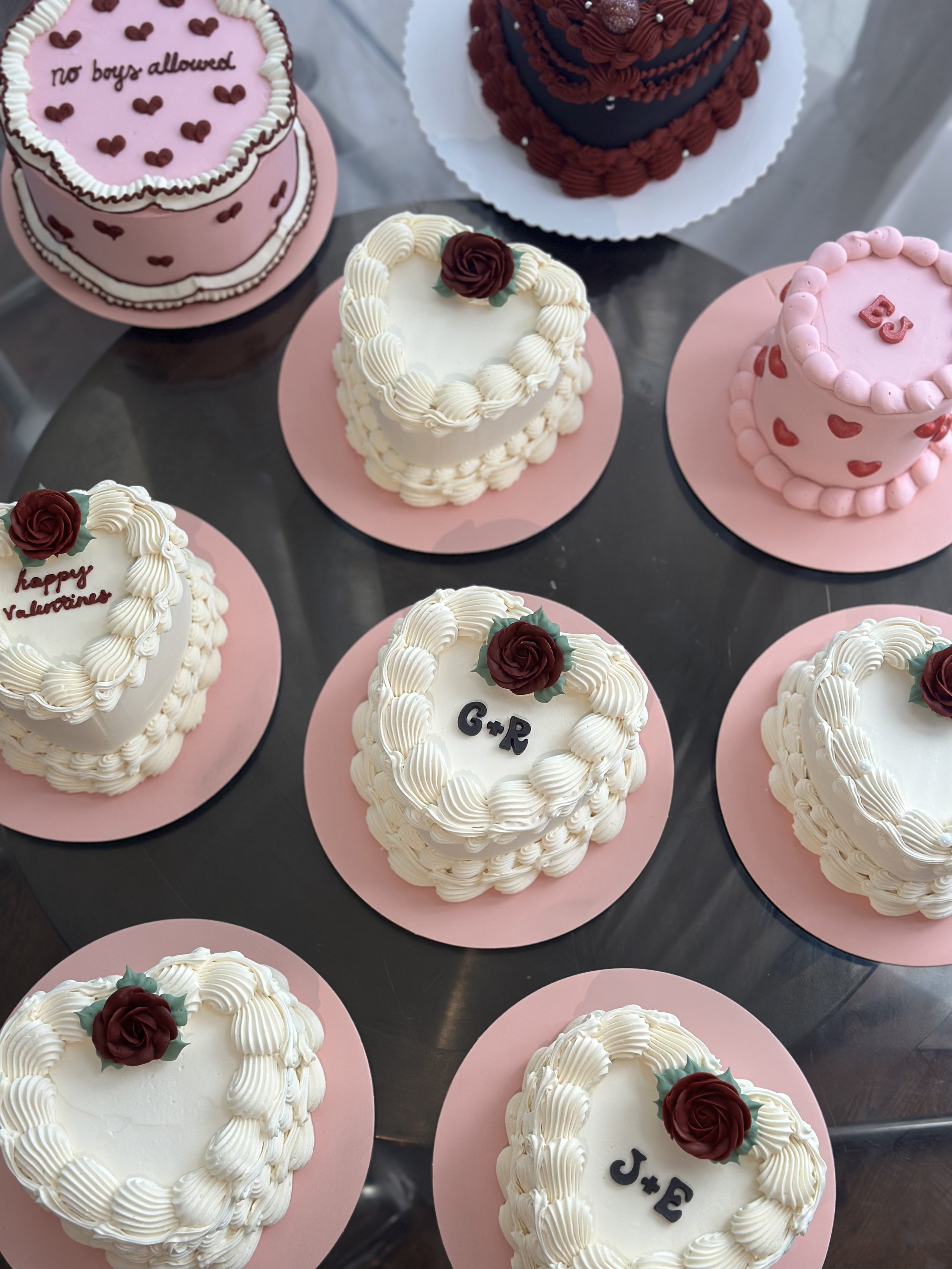 Multiple heart-shaped and round cakes decorated with white, pink, and red icing, some with roses and initials, placed on pink and white bases.