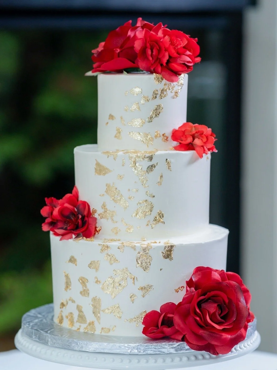 Three-tier white wedding cake decorated with red roses and gold leaf accents.