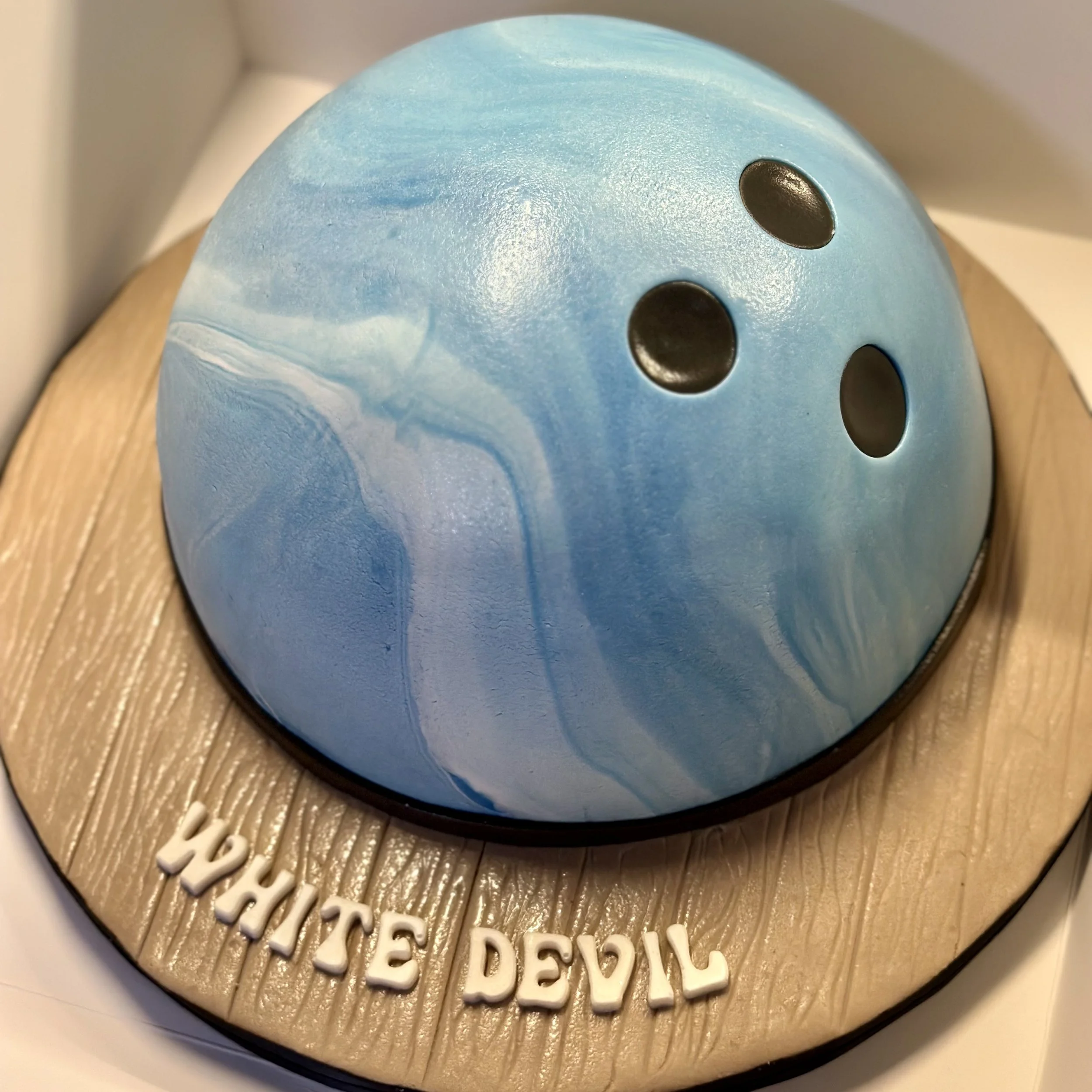 A birthday cake decorated to resemble a bowling ball, with a blue marbled planet on top, placed on a wooden cake board with the words 'white devil' written on it.