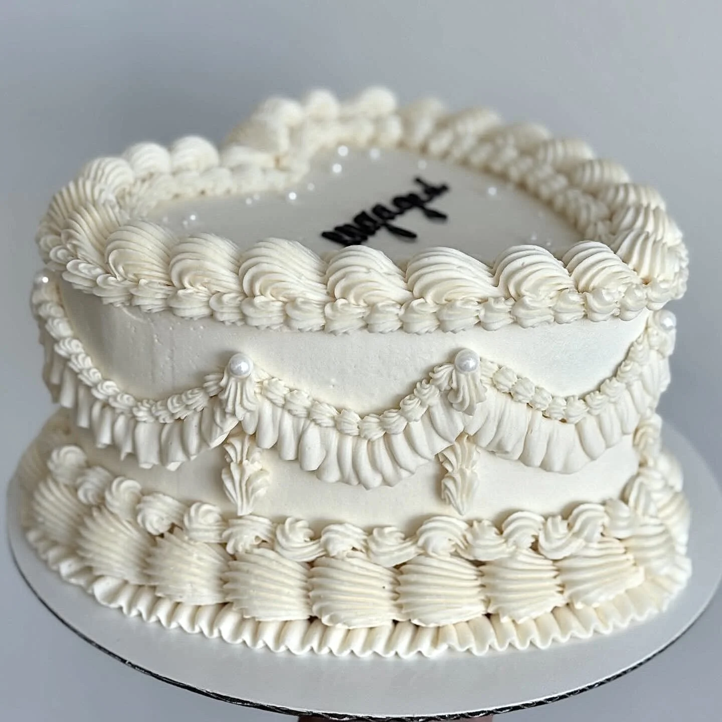 White heart-shaped cake decorated with piped cream designs and pearl accents, with a section of black lettering on the top.