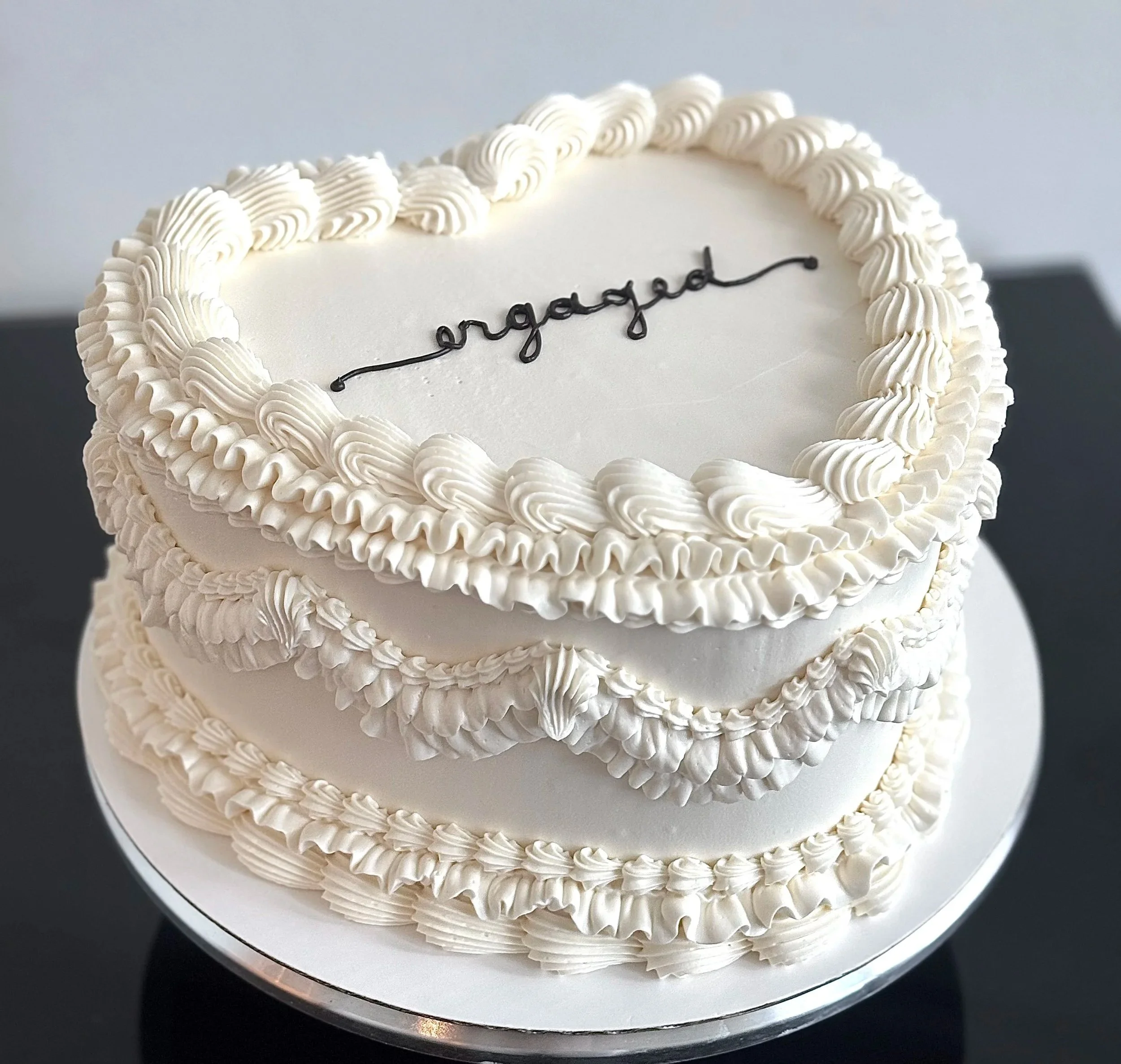 A white heart-shaped cake decorated with piped white frosting and the word 'engaged' written in black icing on top.