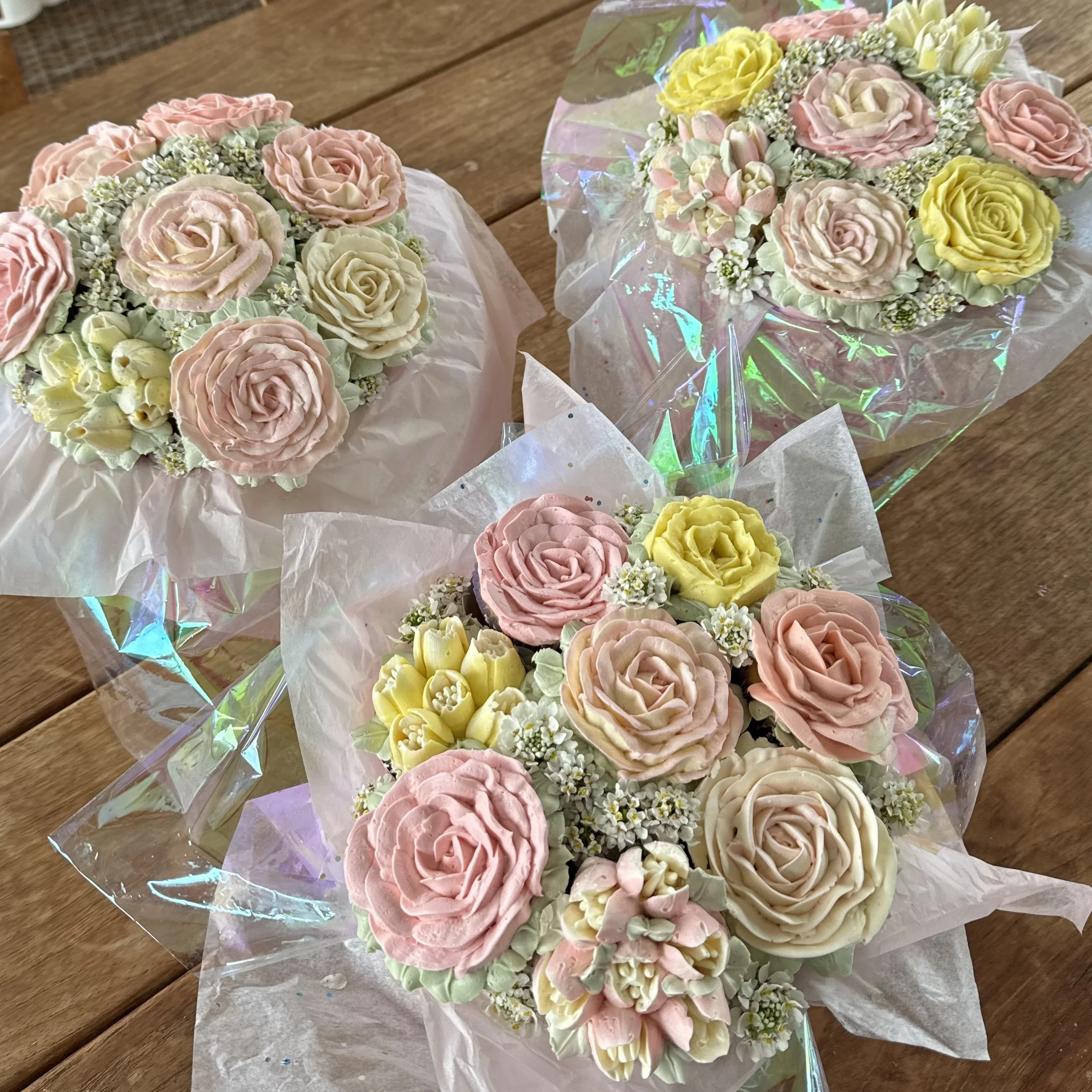 Three bouquets of intricately decorated floral cupcakes with rose designs in pastel pink, yellow, lavender, and white, wrapped in clear and iridescent cellophane, resting on a wooden surface.