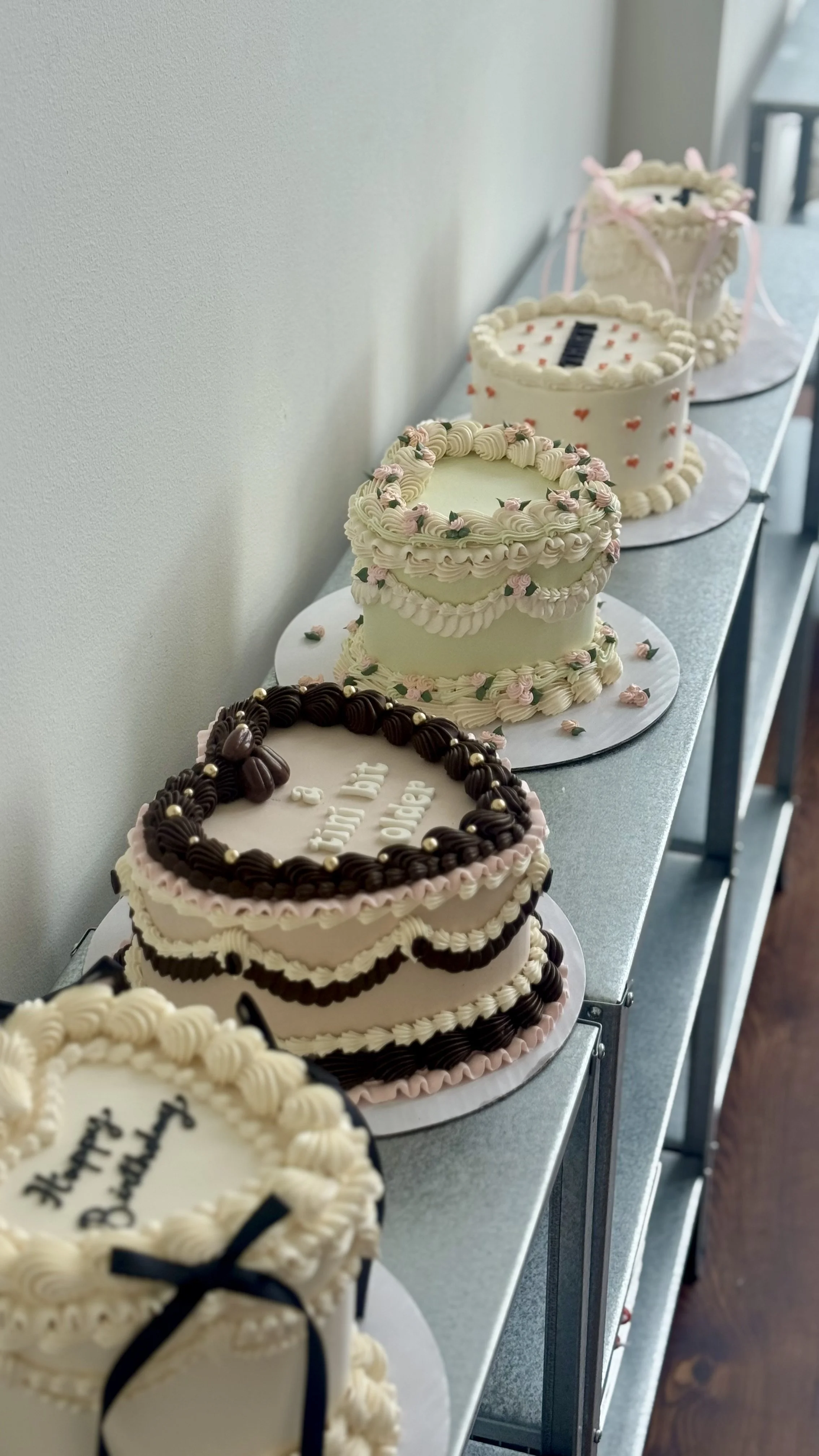A line of decorated birthday cakes on a metal shelf against a wall.