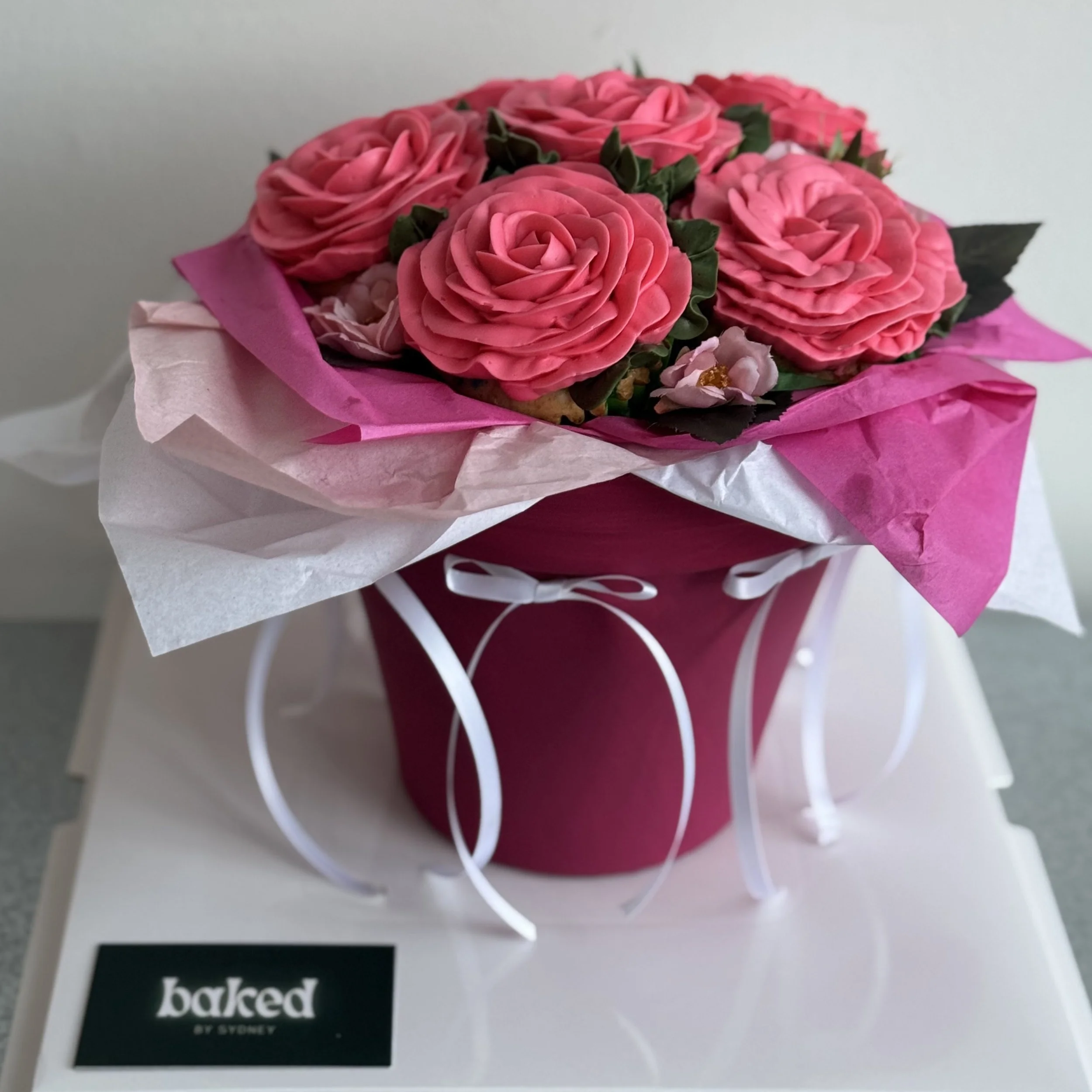 Pink flower bouquet with roses and small flowers in a pink and white wrapping, placed in a small pink pot with white decorative curls, on a white surface.