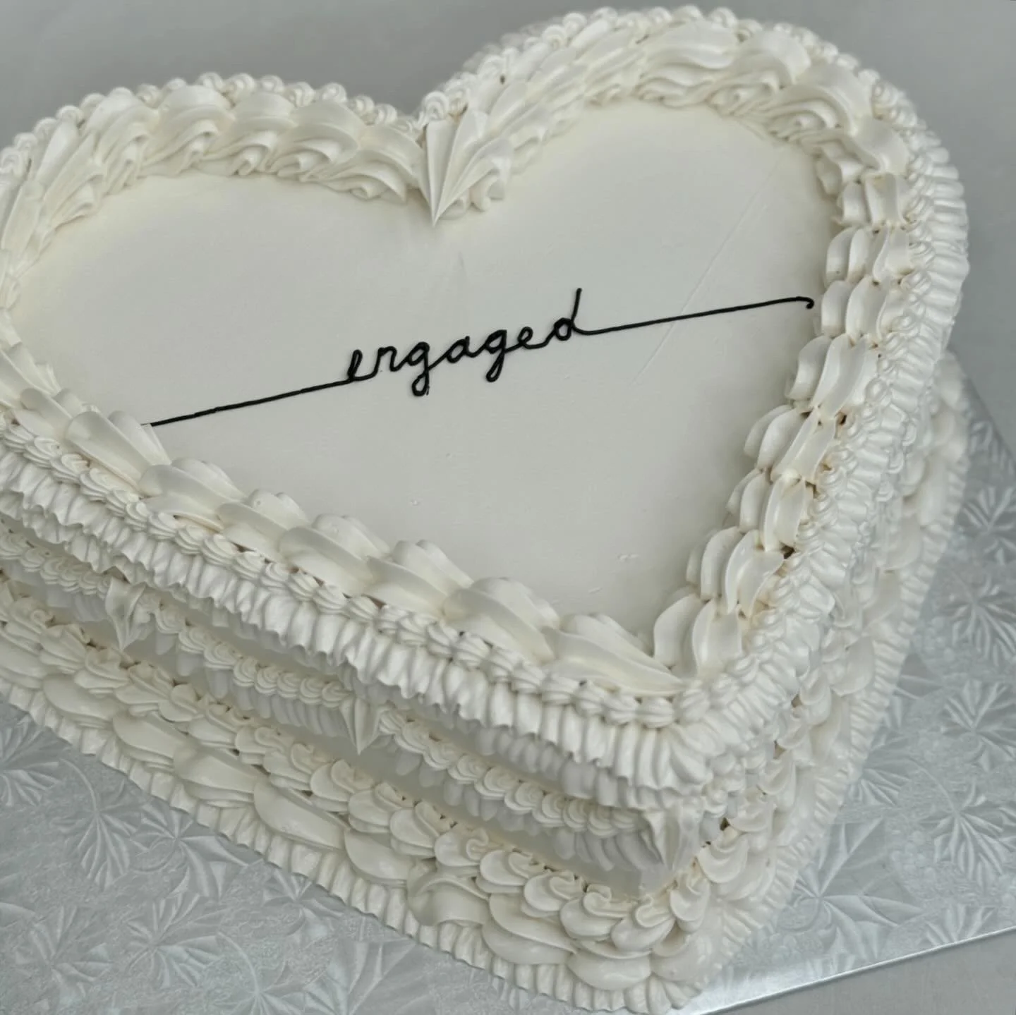 A heart-shaped white cake with decorative white frosting piping and the word 'engaged' written in black cursive on the top.