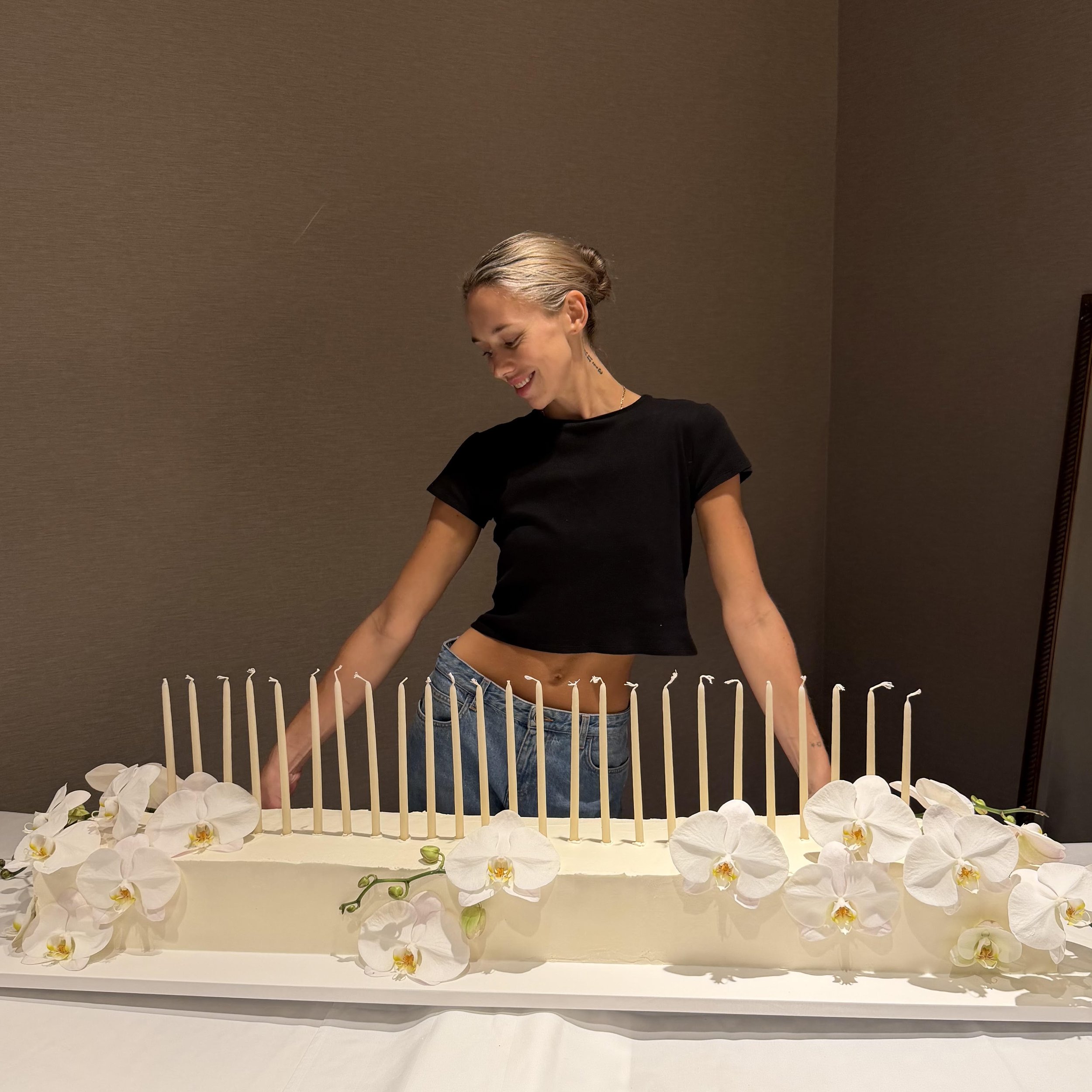 A smiling woman with blonde hair styled back, wearing a black top, stands behind a white table decorated with white orchids and a white fence-like barrier at an indoor event.