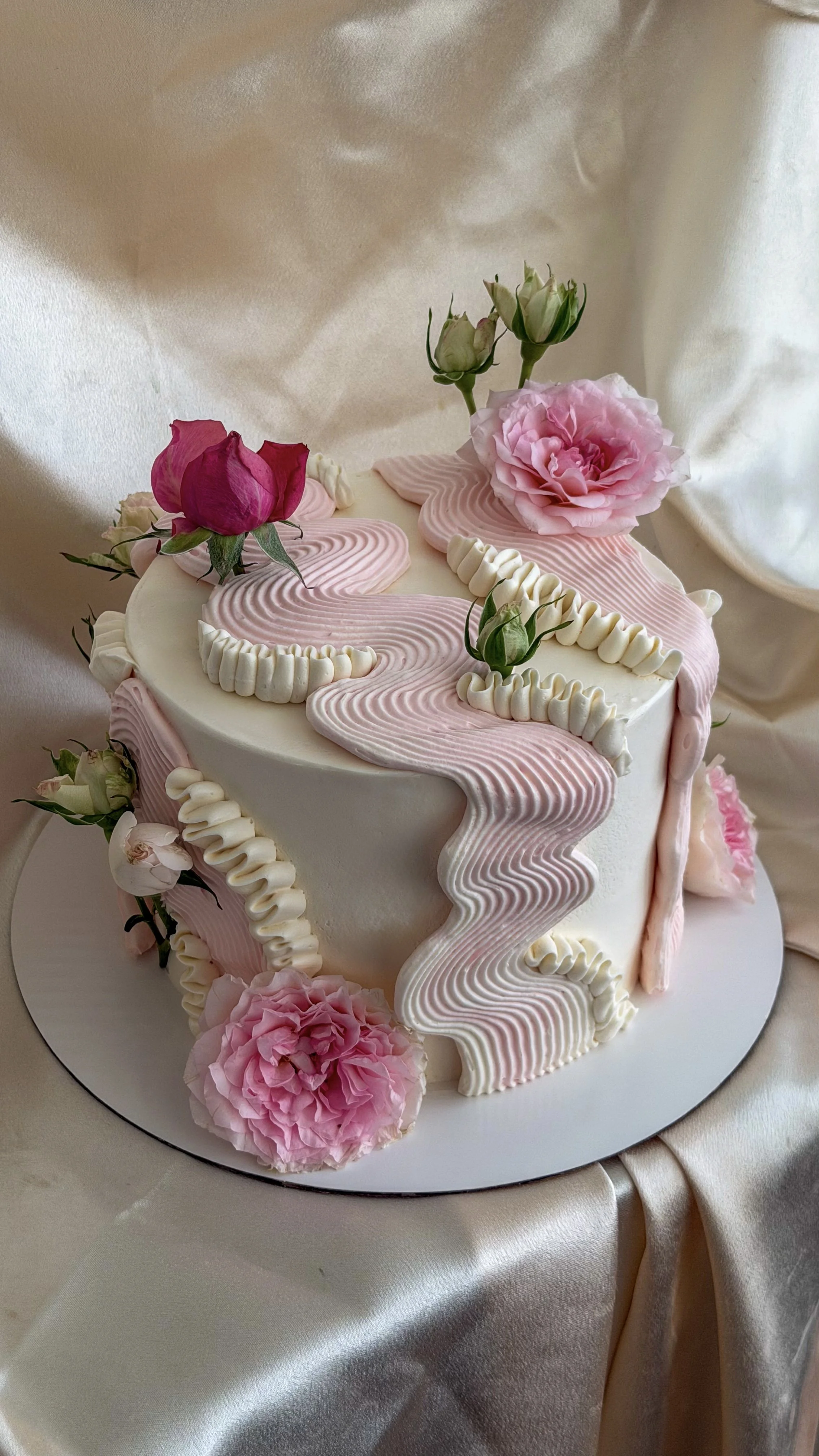 A decorated birthday cake with pink roses and floral accents, topped with pink and white piped icing in a flowing design, on a white cake stand against a satin background.