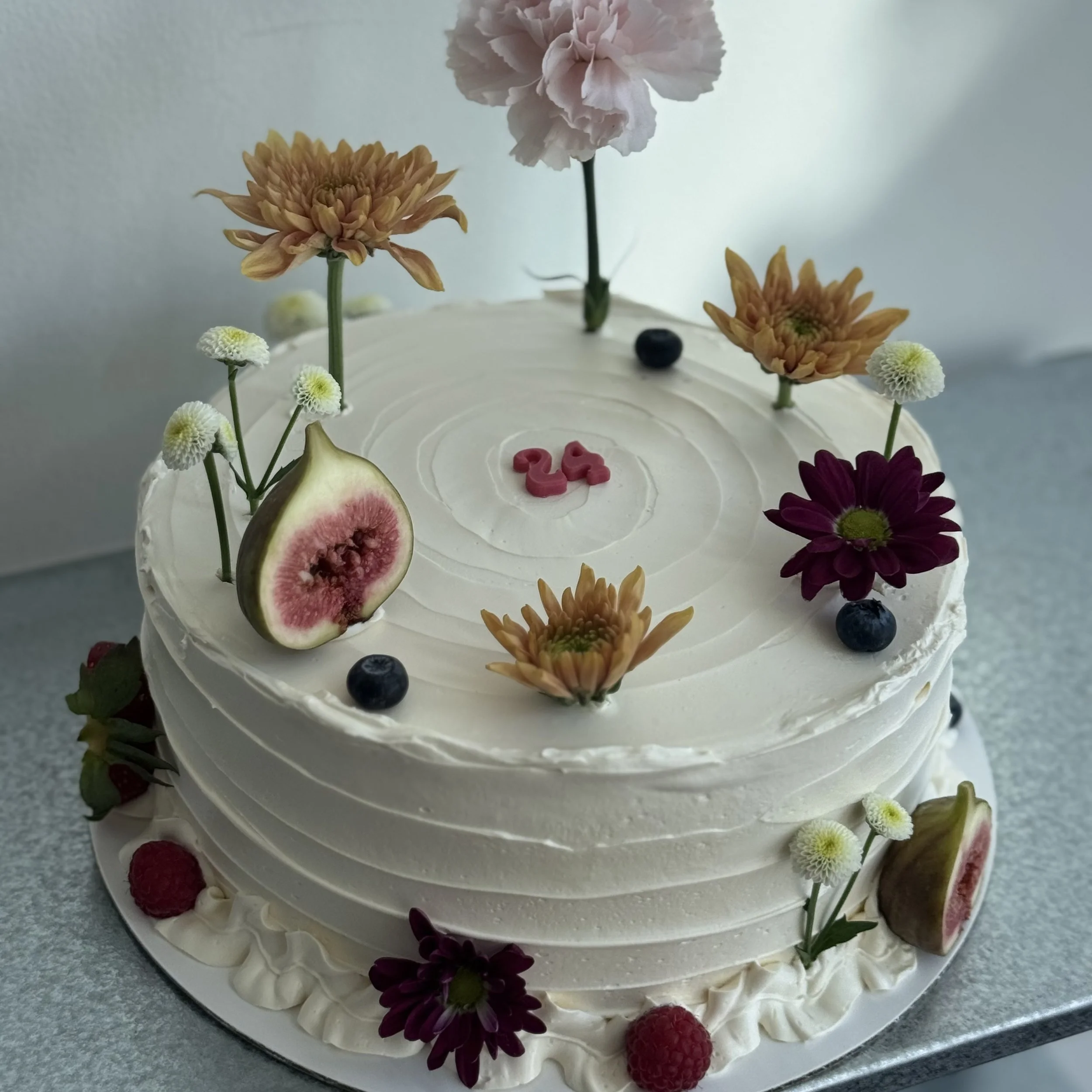 A round white birthday cake decorated with various fresh flowers, figs, blueberries, and raspberries, with a pink number 24 candle on top.
