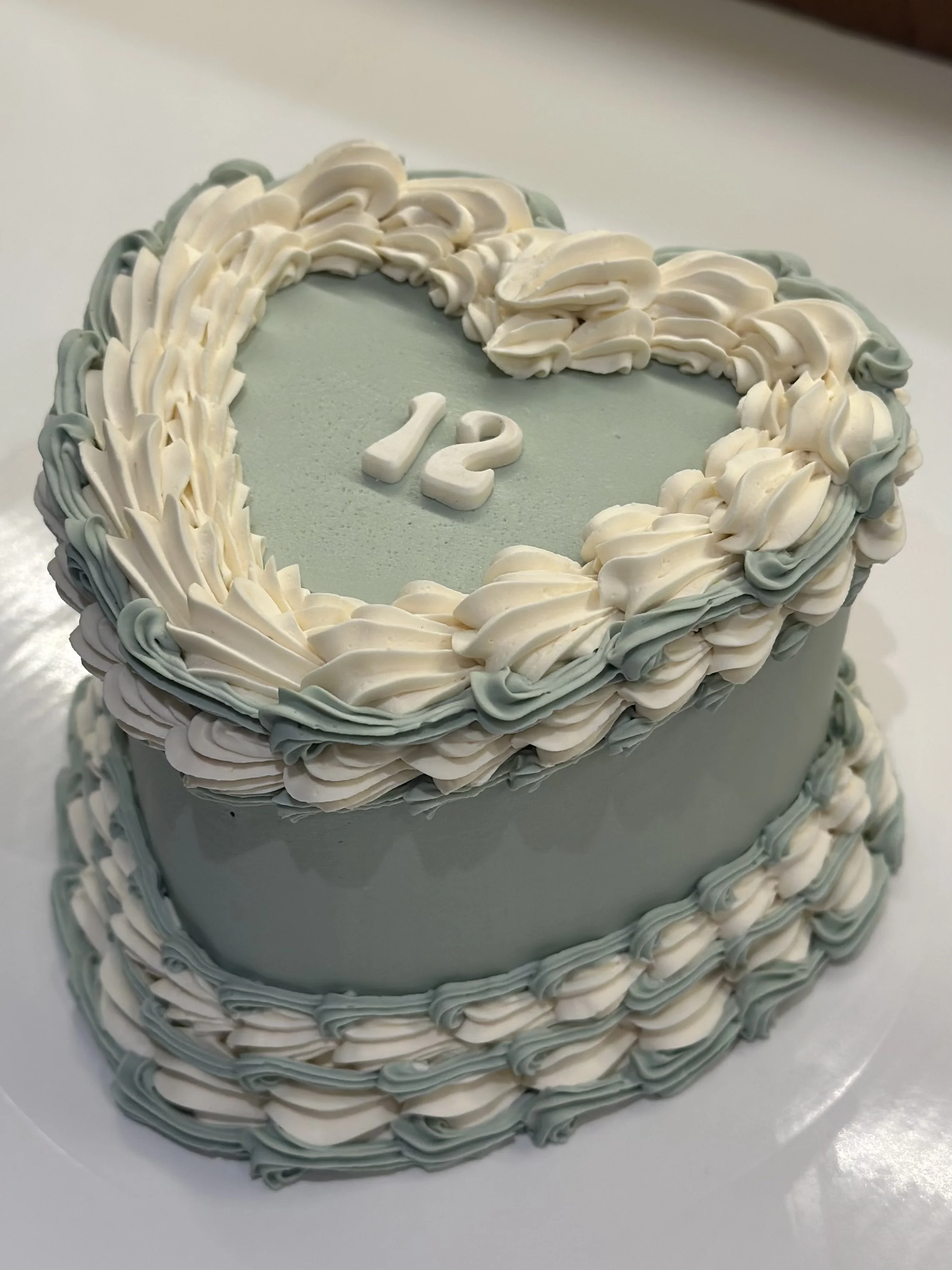 Two-tiered cake with gray and white piped icing, decorated with a heart-shaped border on top, featuring the number '12' in the center.