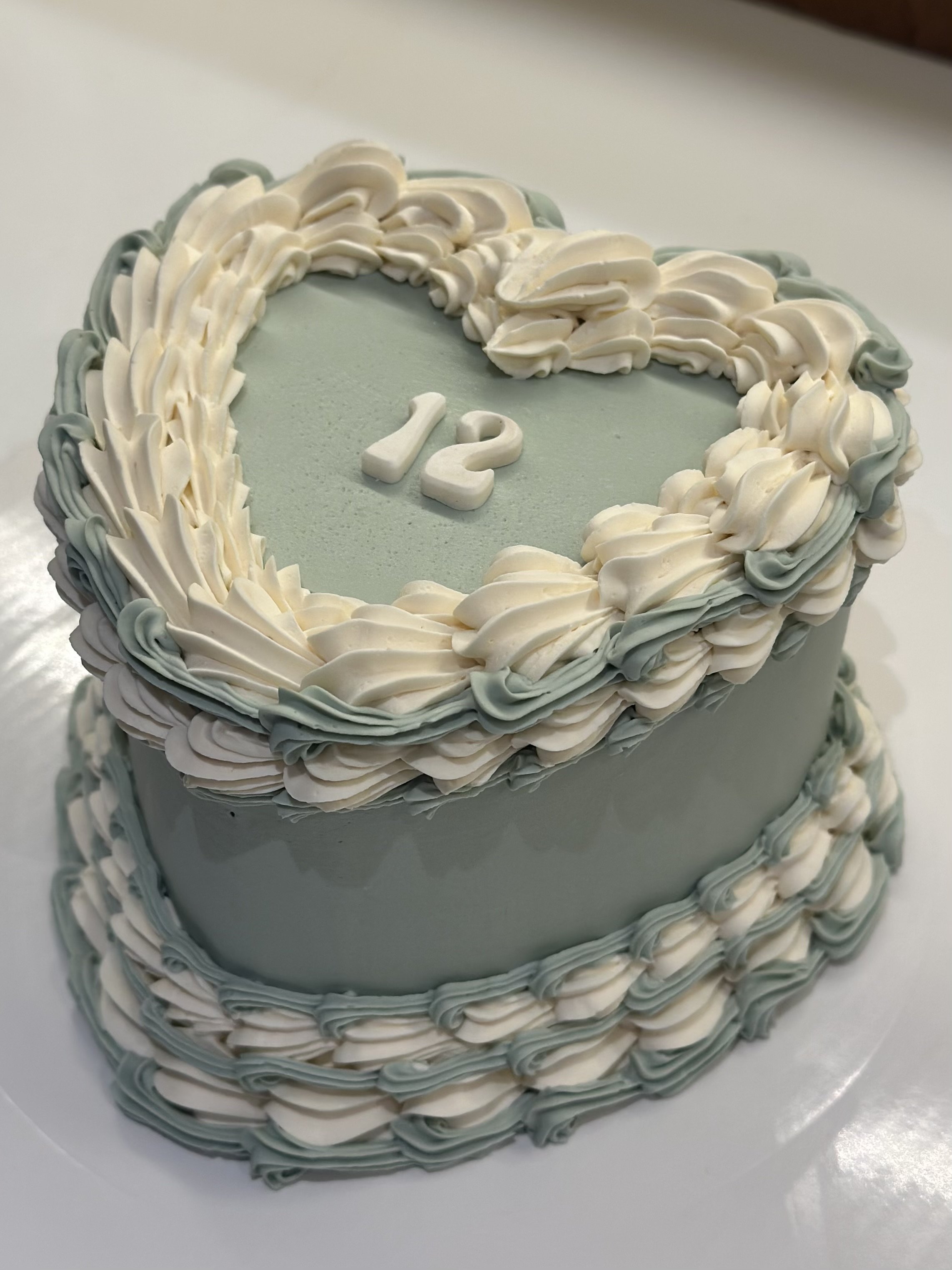 Two-tiered cake with gray and white piped icing, decorated with a heart-shaped border on top, featuring the number '12' in the center.