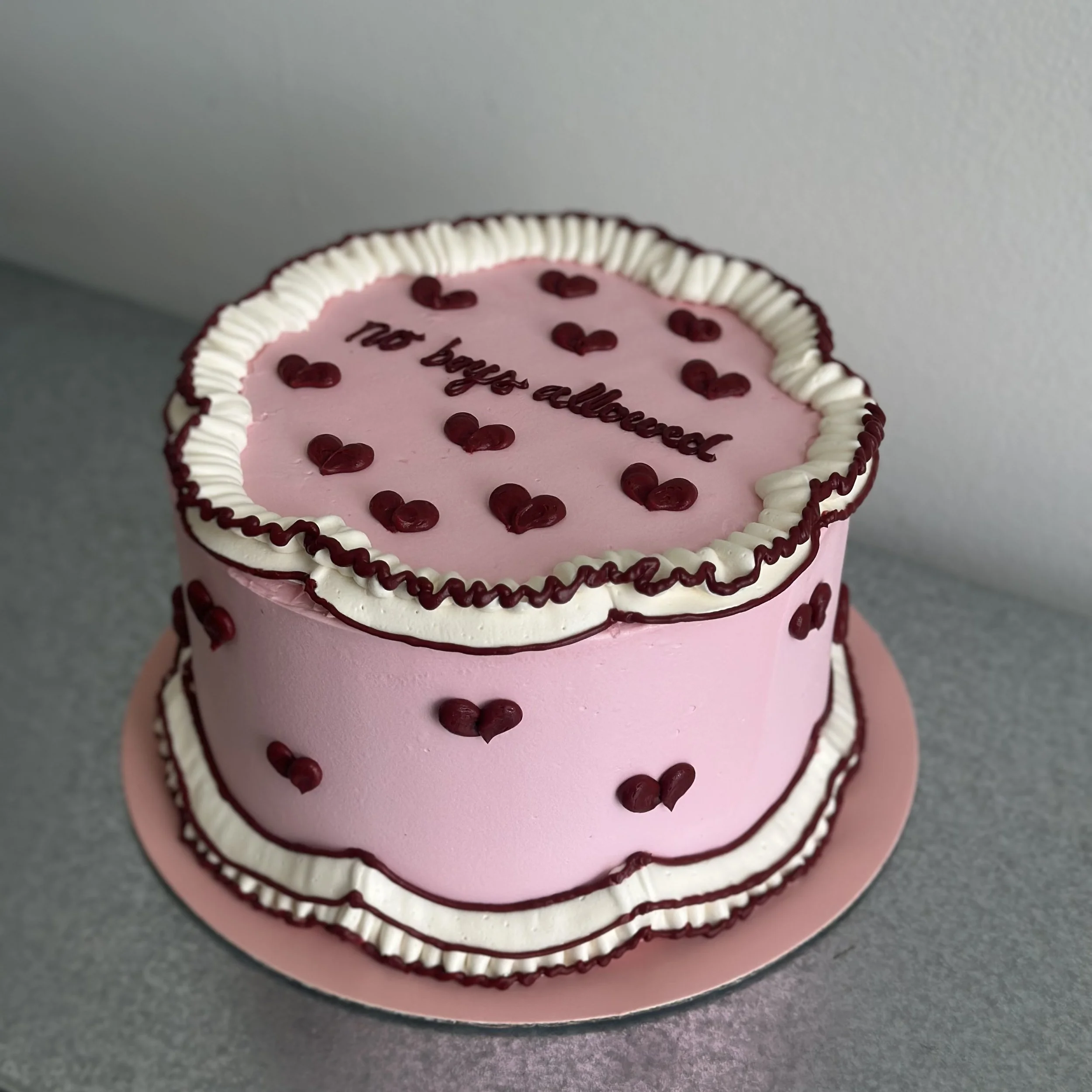 A pink two-tiered cake decorated with red heart-shaped sprinkles and white and dark red frosting borders. The top of the cake has the message 'No bugs allowed' written in dark red icing.