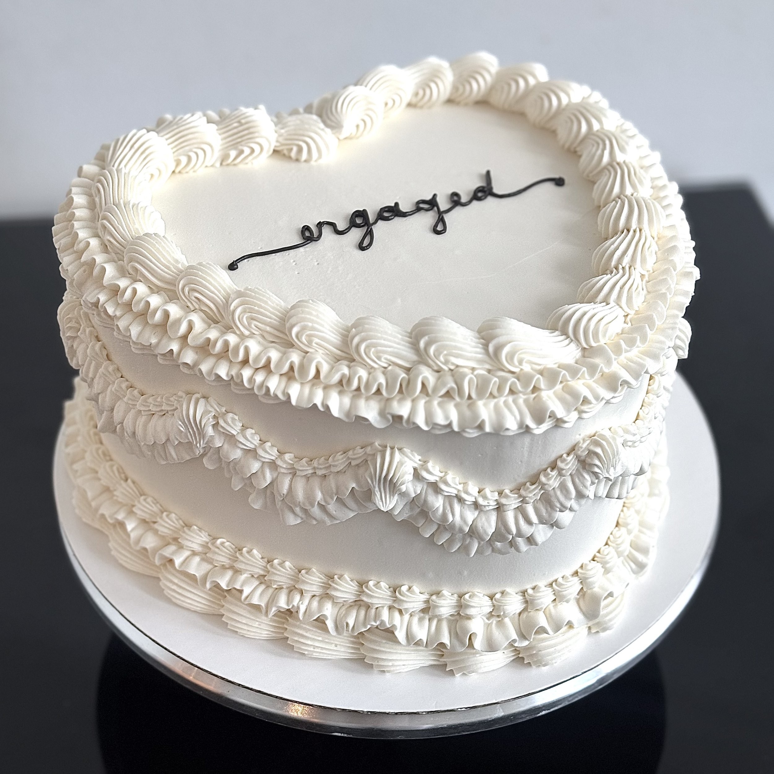 A white heart-shaped cake with intricate piped borders and the word 'engaged' written in black icing on the top.