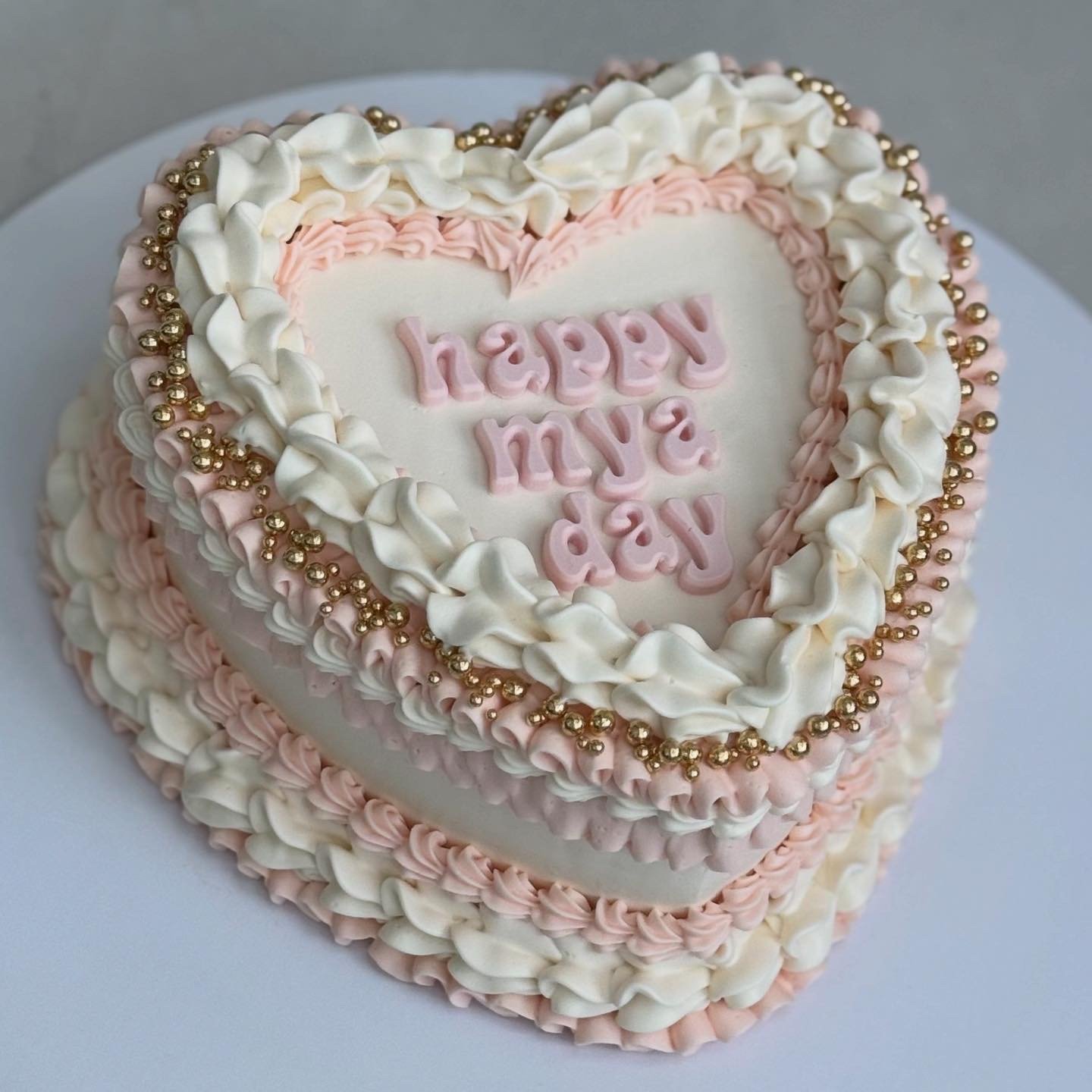 A heart-shaped celebration cake decorated with white and pink frosting, gold beads, and pink lettering that says "Happy May Day".