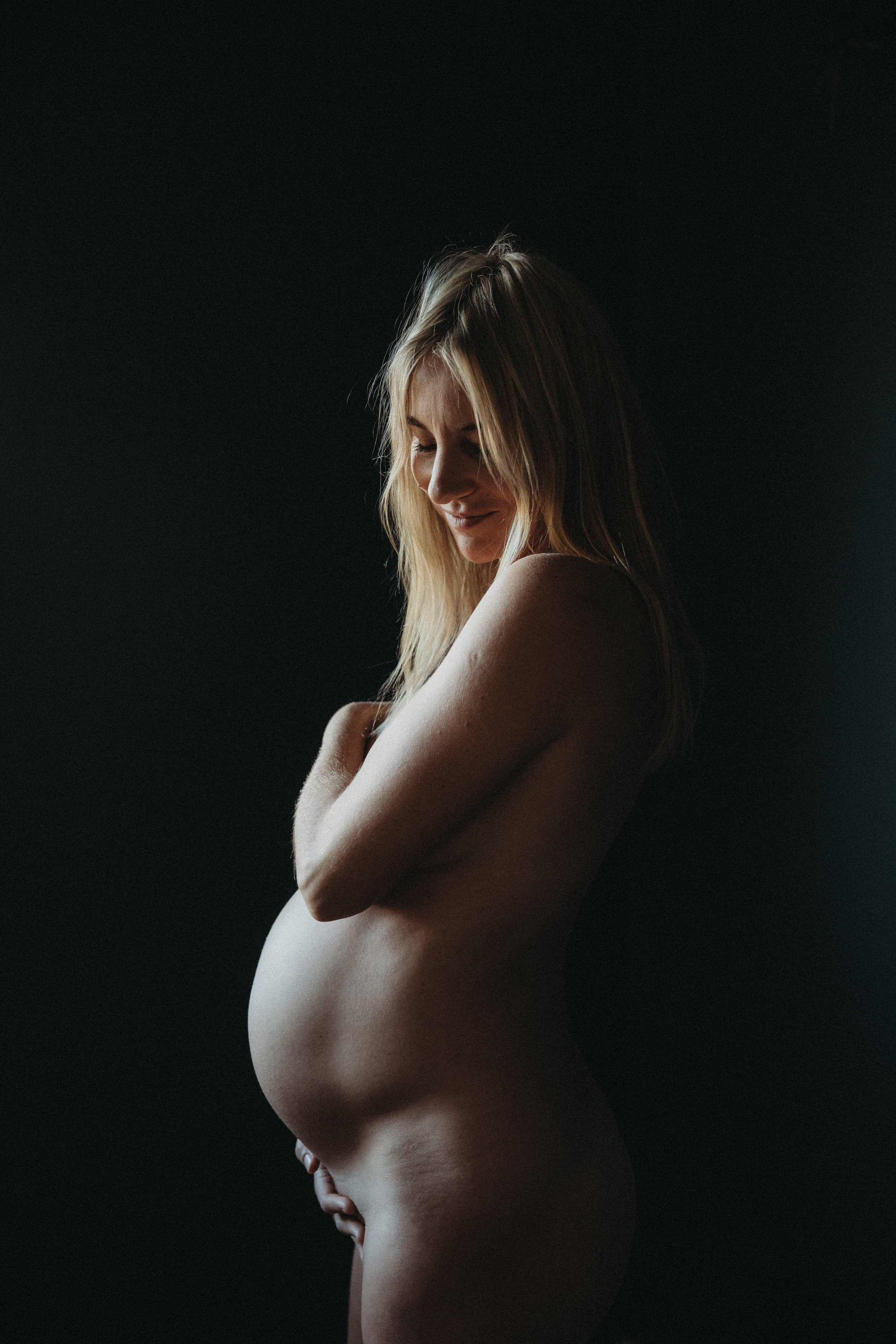 Pregnant woman with long blonde hair, standing and looking down, set against a dark background.