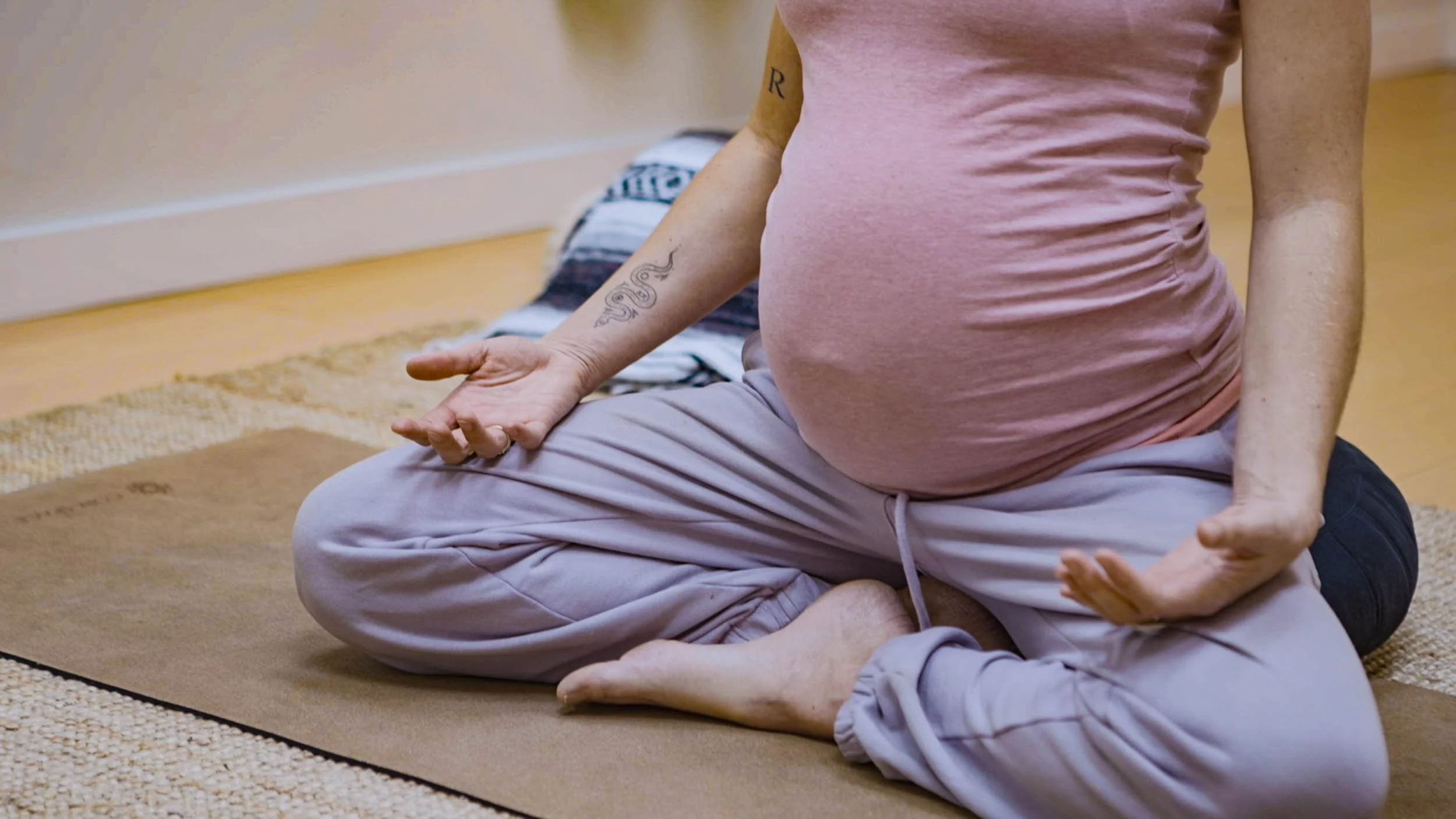Pregnant person sitting cross-legged on a yoga mat, meditating with hands in mudra position.