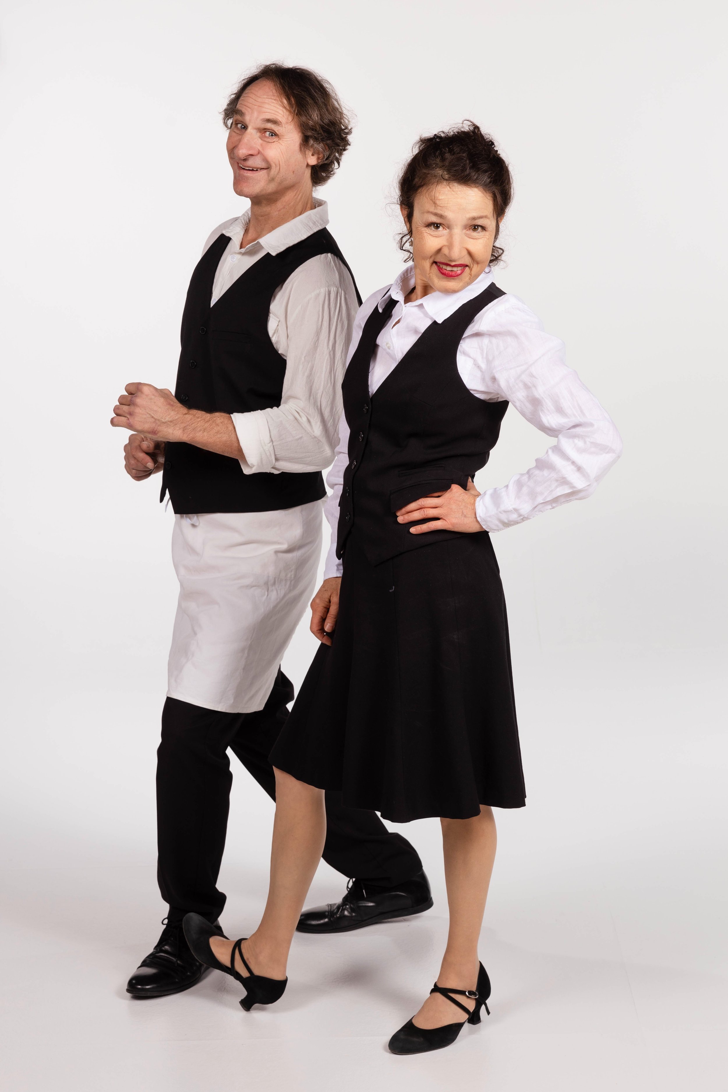 Due camerieri comici sorridenti in abiti da lavoro eleganti, camicia bianca e gilet nero, su sfondo bianco.