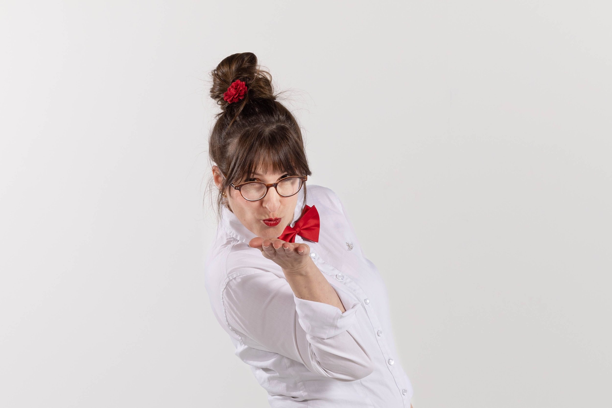 Donna che soffia un bacio, indossando occhiali, camicia bianca e papillon rosso, capelli raccolti con fiore rosso.