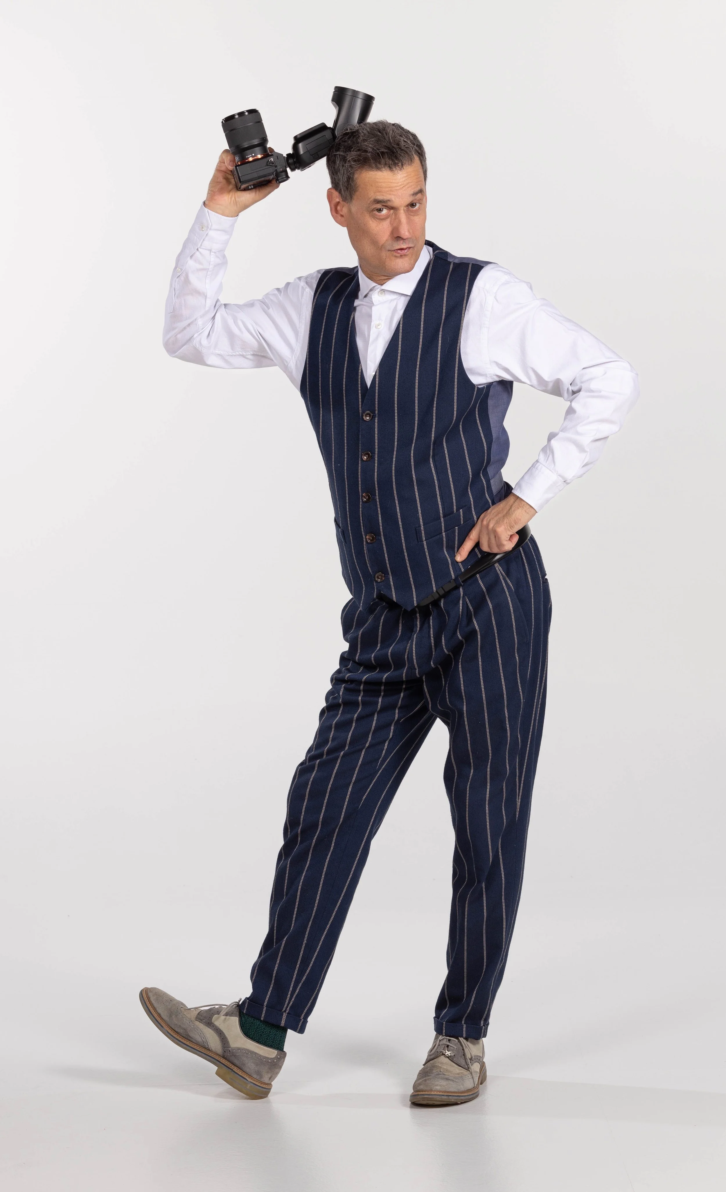 Uomo in abito gessato blu e camicia bianca, posa con una fotocamera sopra la testa su sfondo bianco.