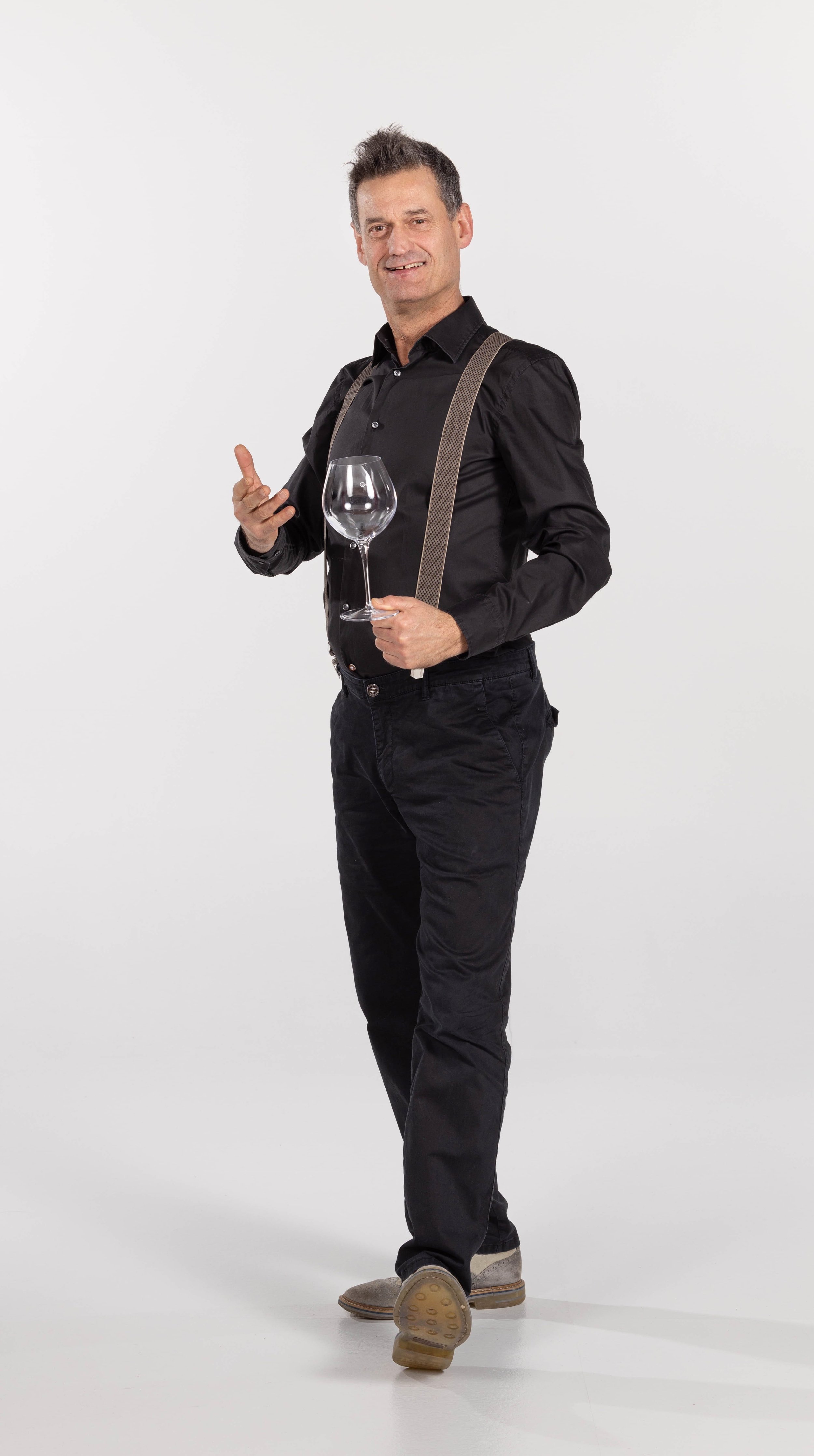Uomo in piedi con un grande bicchiere da vino in mano, indossa camicia nera e bretelle, sfondo chiaro.