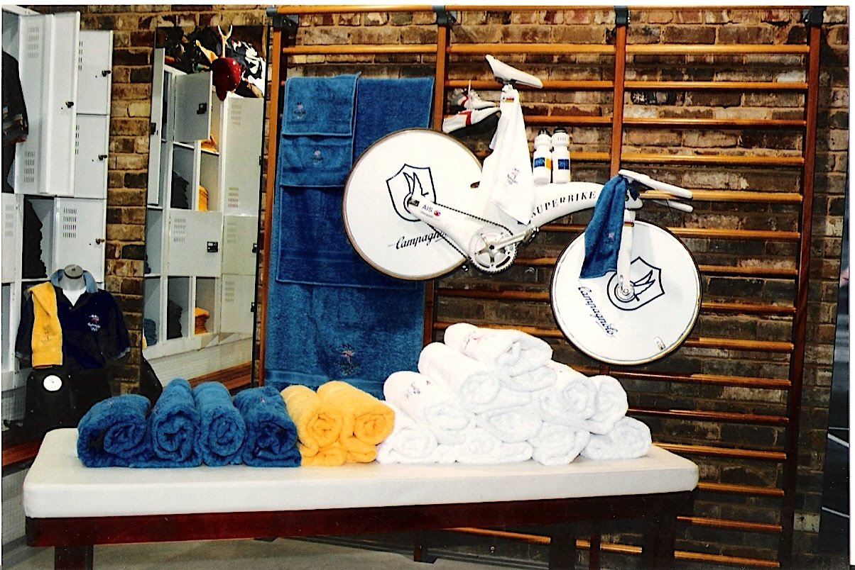 Sports-themed display with rolled towels in blue, yellow, and white on a table, a bicycle with water bottles and towels draped over it, against a brick wall with wooden gym equipment and lockers in the background.