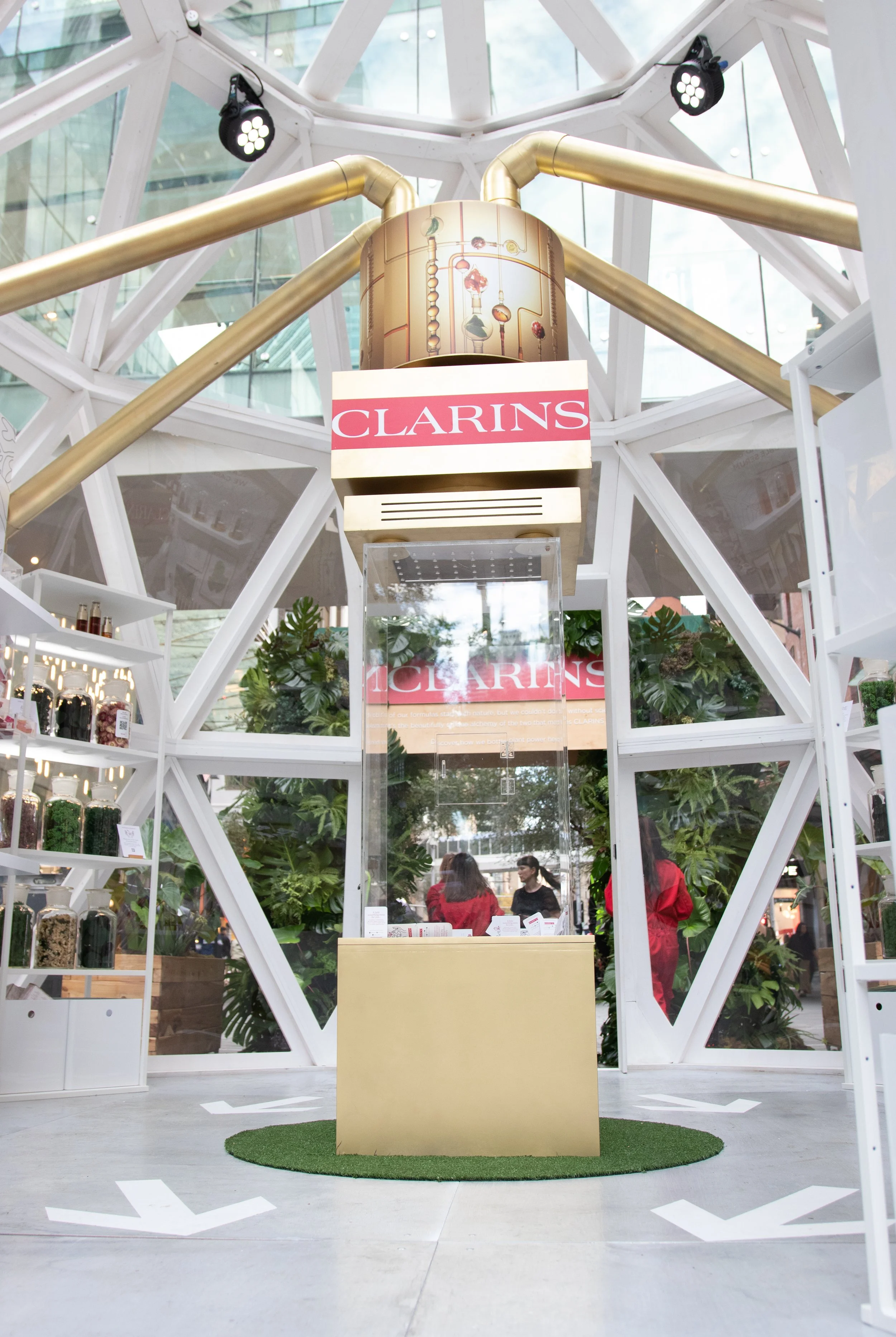 Popup Shop with a Clarins sign, surrounded by plants, with a gold counter in front and arrows on the floor, at an indoor event.
