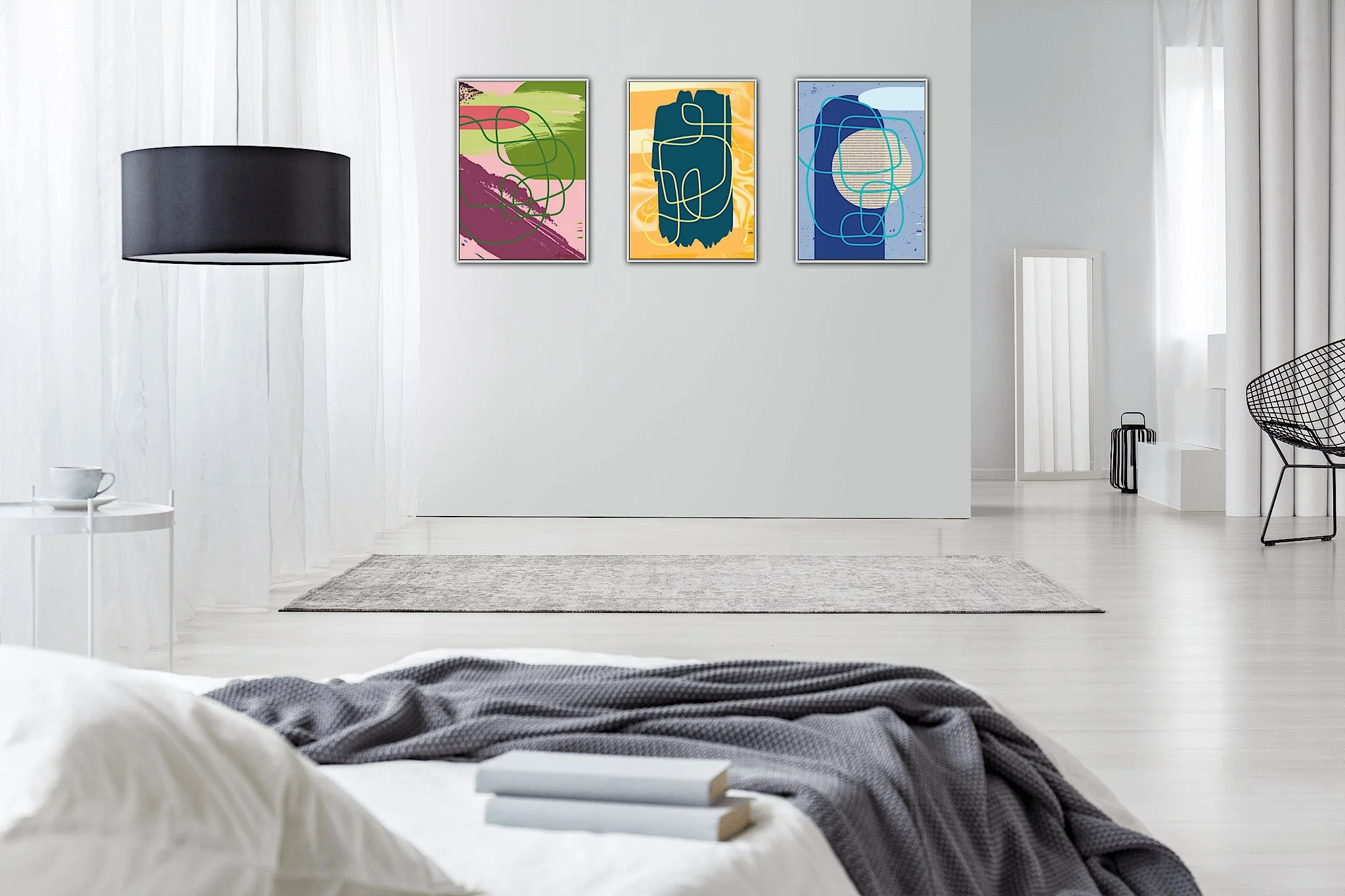 Minimalist bedroom with white walls and floor, black pendant light, three colorful abstract artworks on the wall, a white side table with a cup, a white bed with gray blanket and books, black chair with wireframe design, and a mirror leaning against 