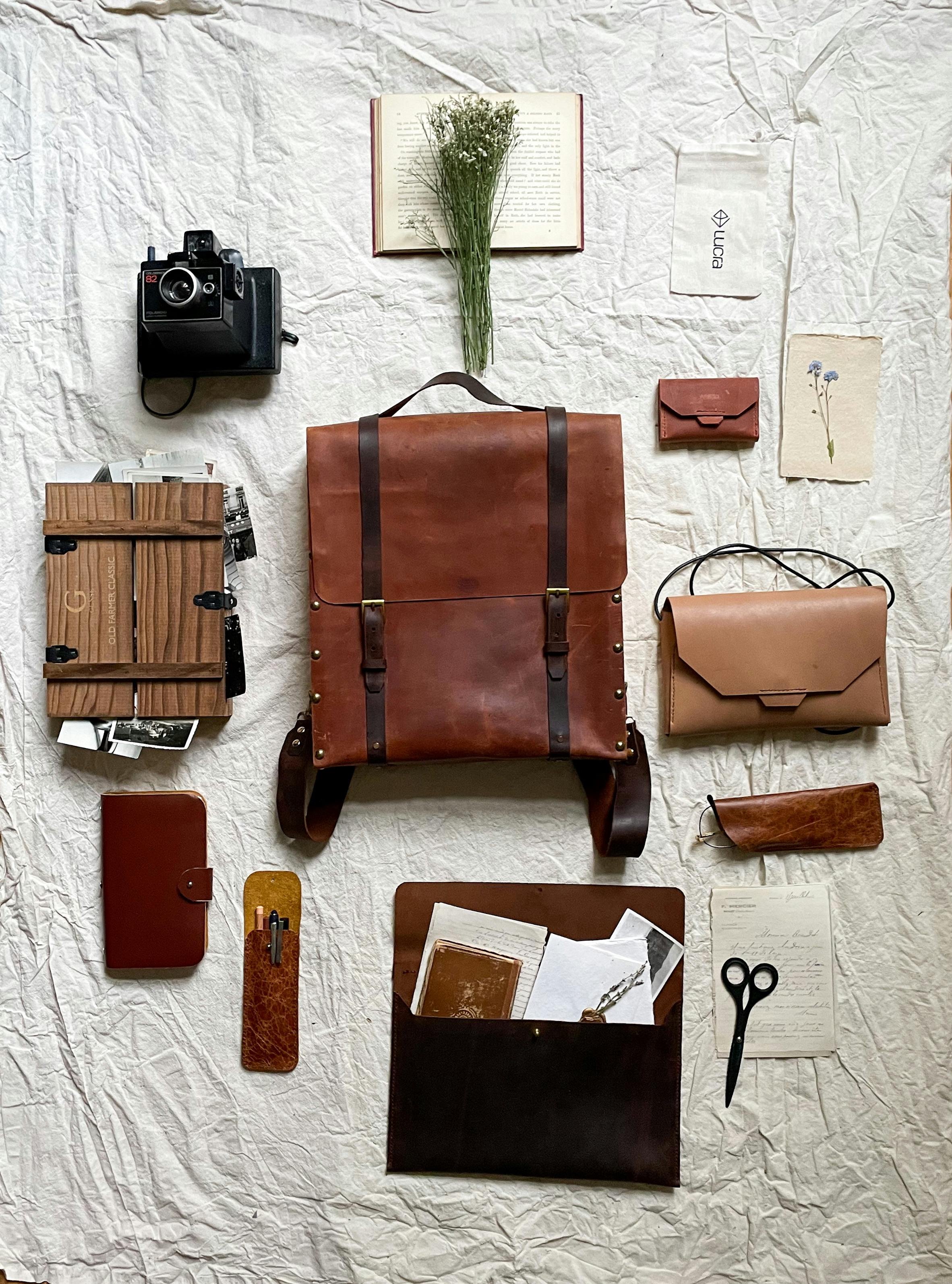 Flat lay of assorted brown leather and wooden items, a camera, books, a notebook, writable papers, scissors, a small wallet, a pouch, a flower bouquet, and a greeting card on crumpled white paper background.