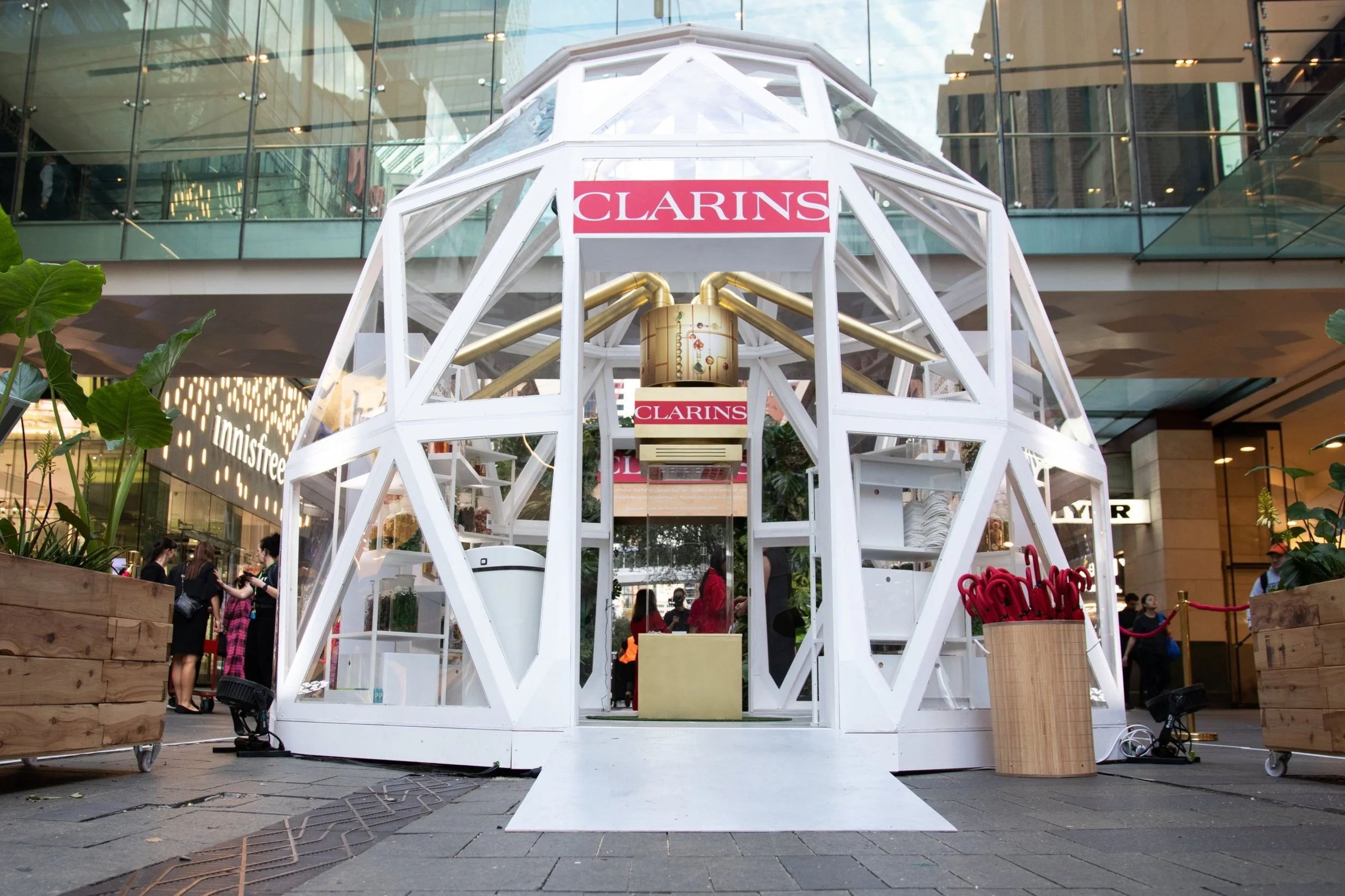 A modern, geodesic white structure in a shopping mall, with Clarins branding, contains greenery and promotional items, surrounded by shoppers and stores.