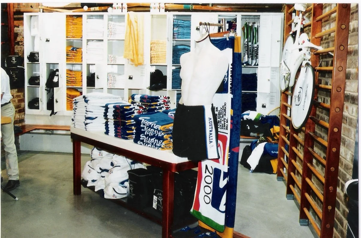Display of sports merchandise including stacked towels, a mannequin torso with Australian rugby shorts, scarves, and bags, with lockers and a wooden gymnastic wall in the background.