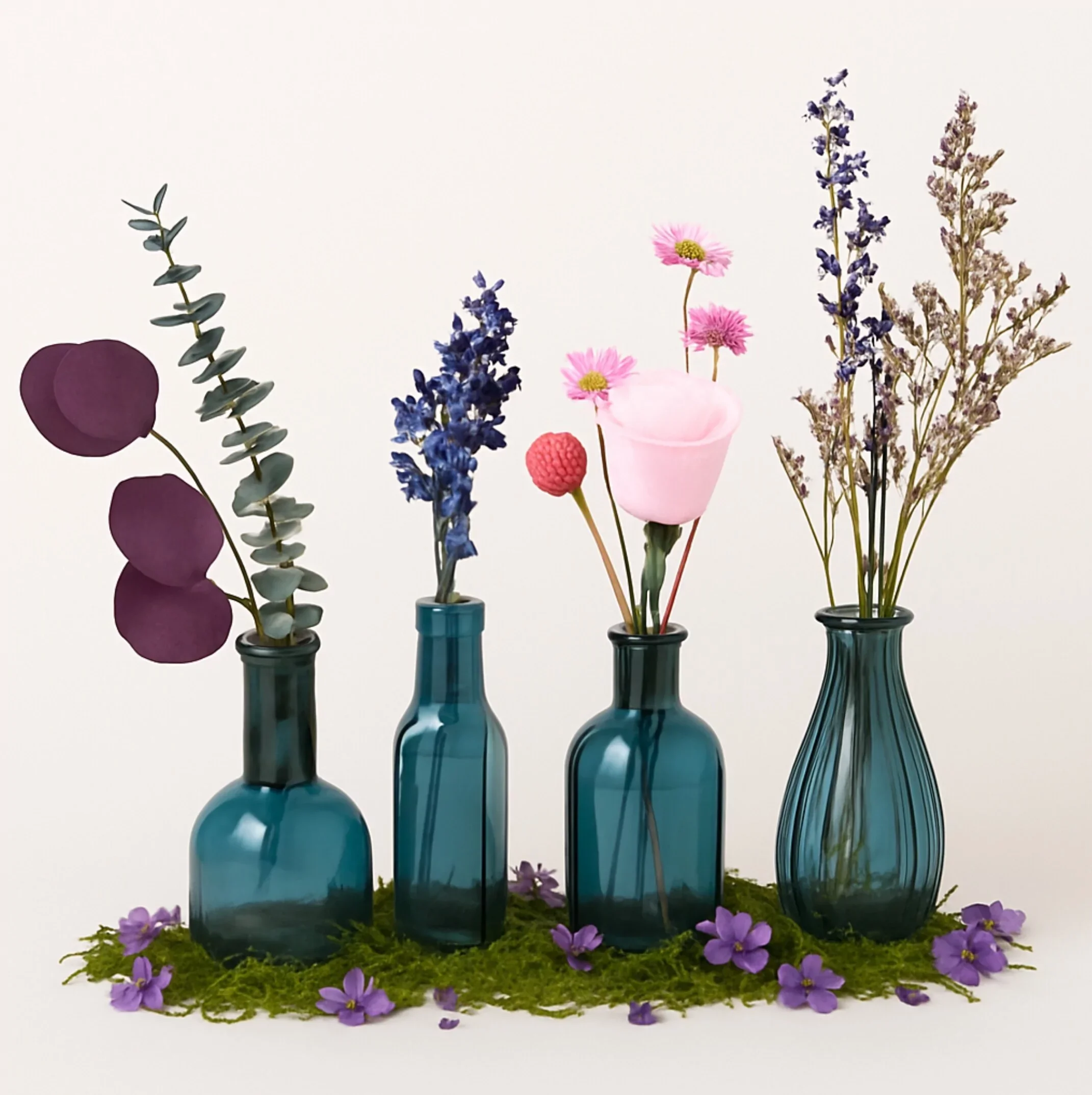 Four teal glass vases with different types of flowers, decorative purple flowers and green moss at the base, against a plain white background.