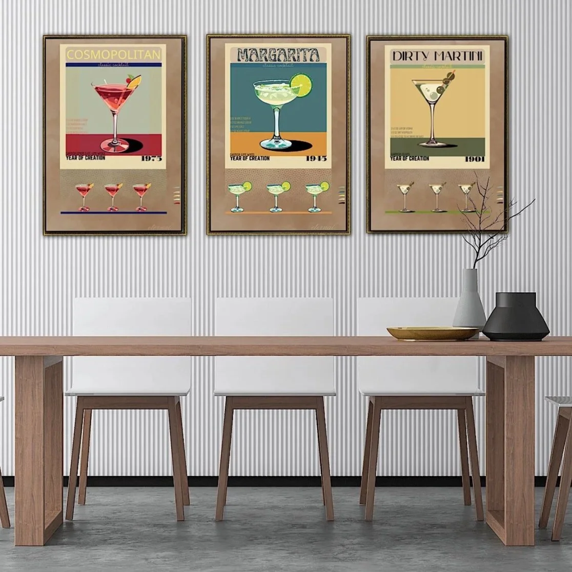 Three framed posters of cocktails hanging on a textured wall above a wooden dining table with white chairs and decorative vases.