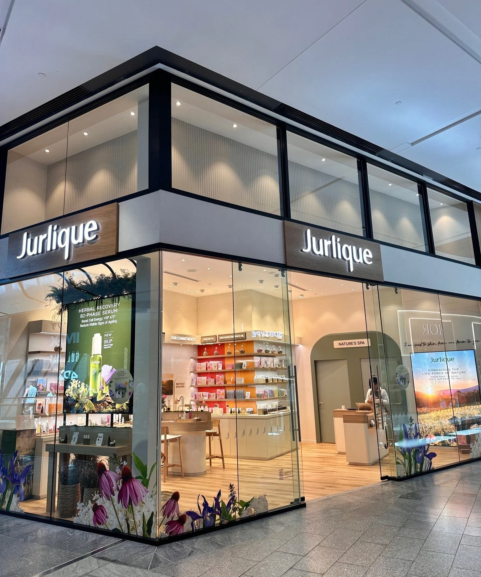 Storefront of Jurlique with large glass windows, displaying skincare products inside, floral decorations outside, and illuminated sign reading 'Jurlique'.