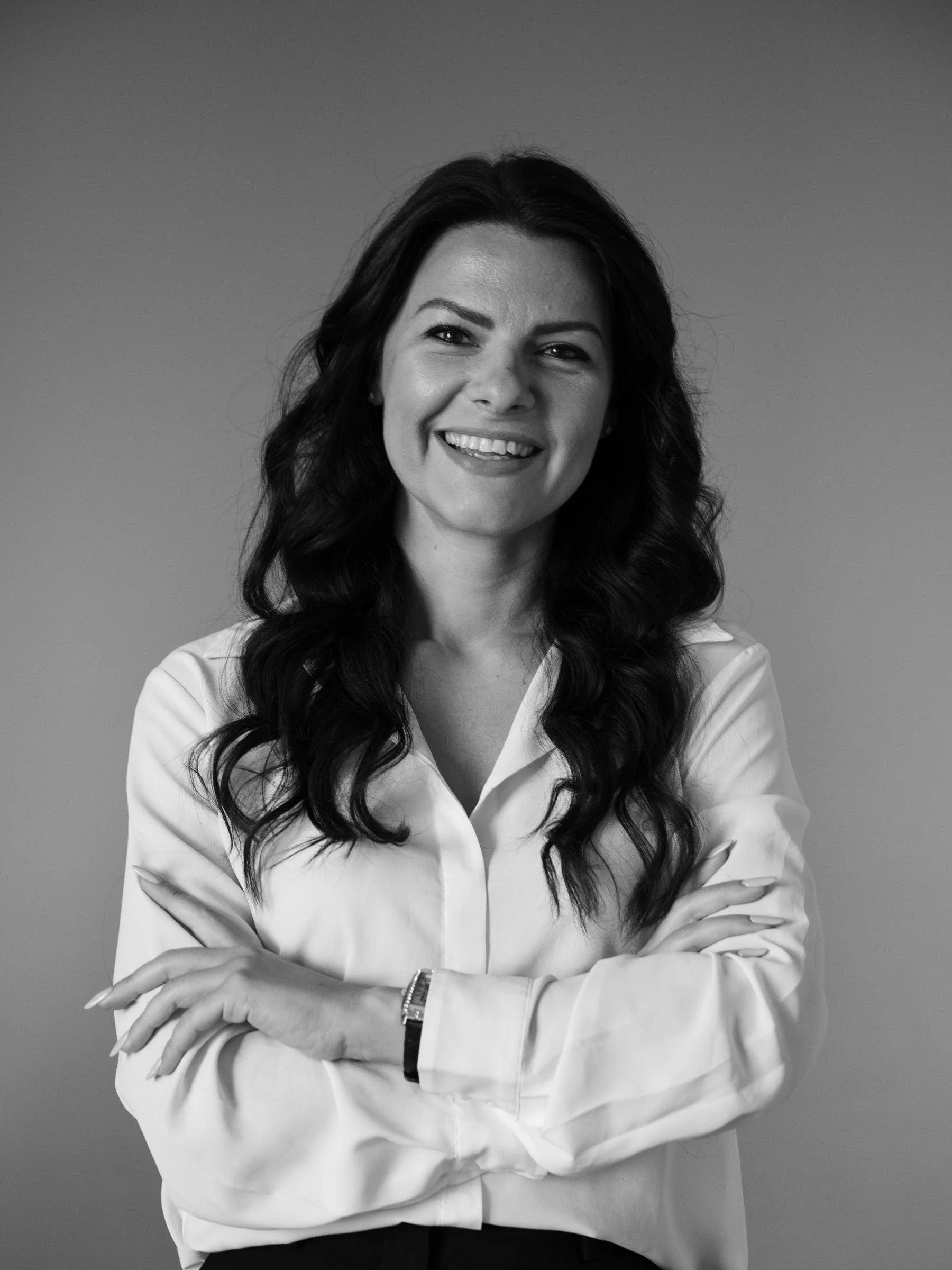 Black and white portrait of a woman smiling, with long dark hair, wearing a white shirt and a watch, arms crossed.