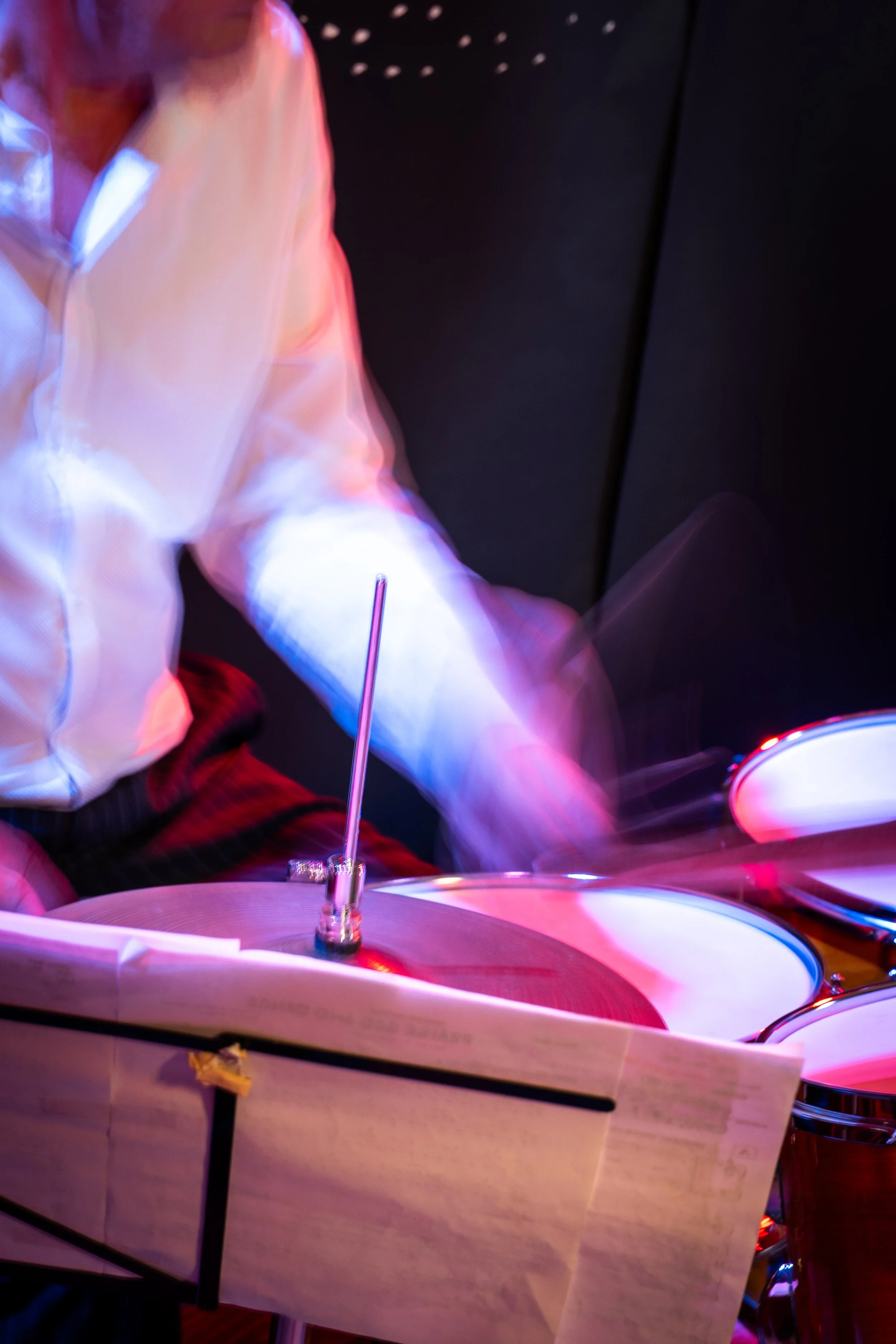 A person playing a drum kit, wearing a white jacket with pink and purple lighting effects, in a dark setting. Sheet music is visible in the foreground.