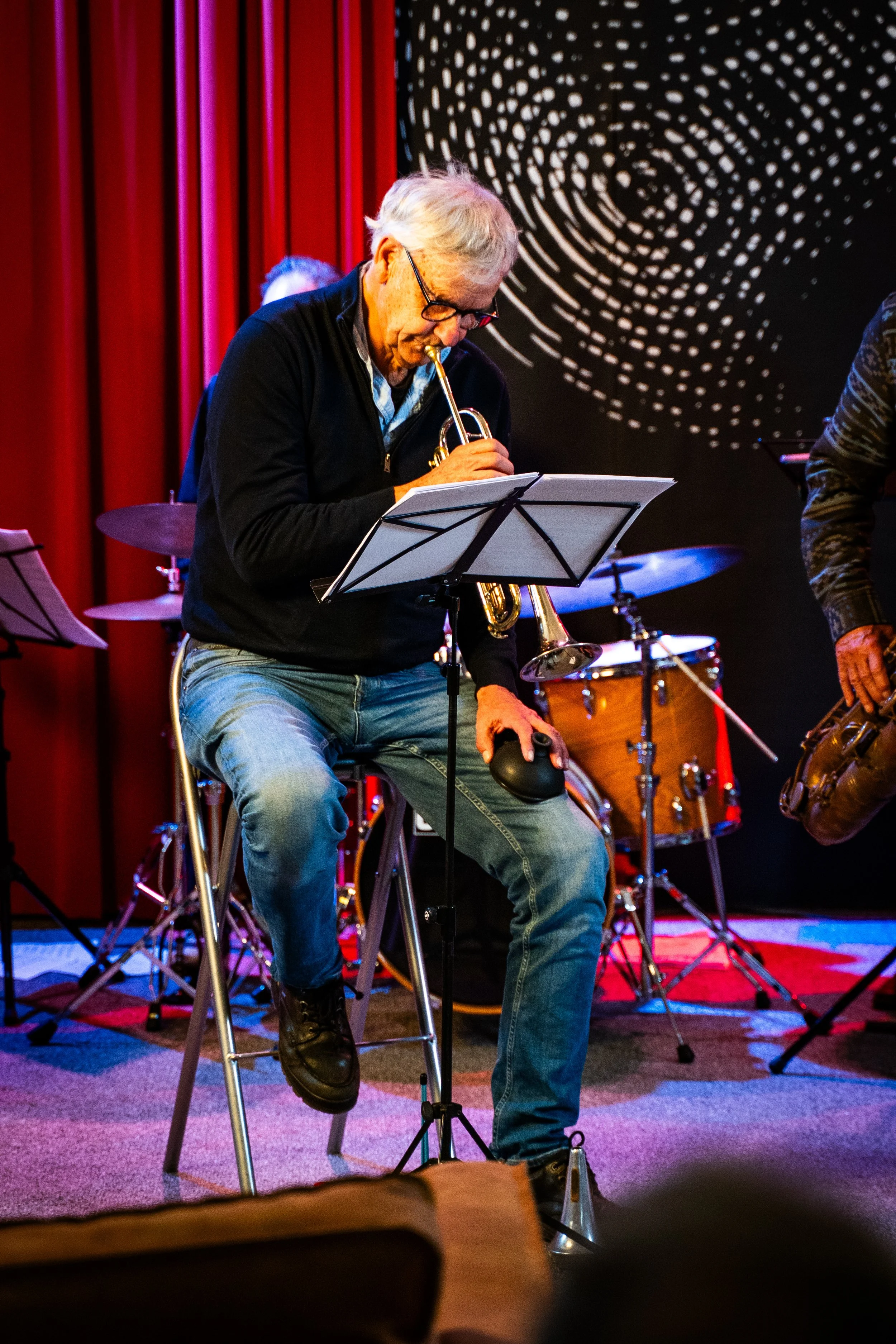 An elderly man with gray hair and glasses playing a trumpet, seated on a chair with a music stand in front of him, during a live music performance in a venue with red curtains and black walls with circular patterns.